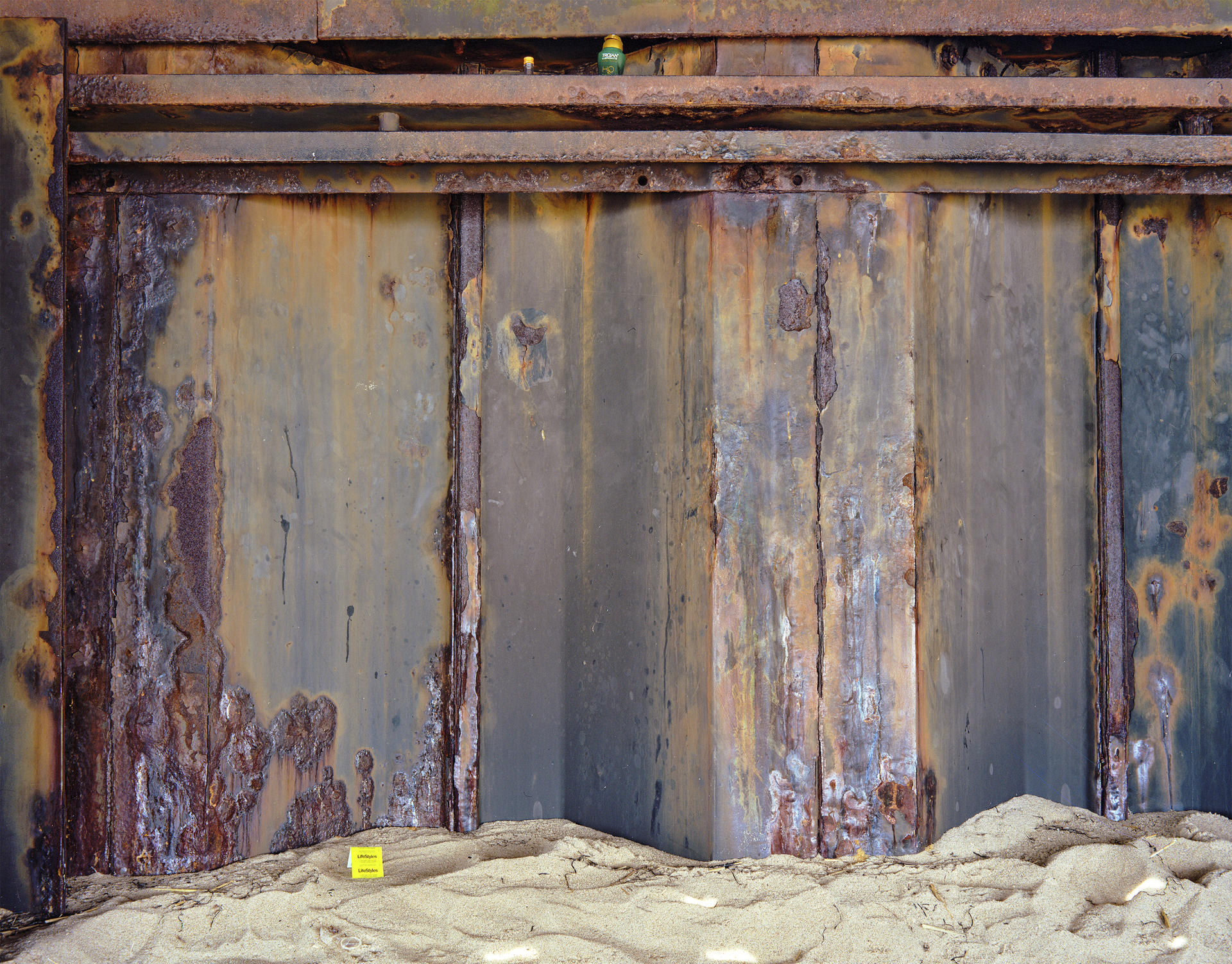 a landscape photograph showing a salt rusted wall with a magnum condom wrapper in the sand. 