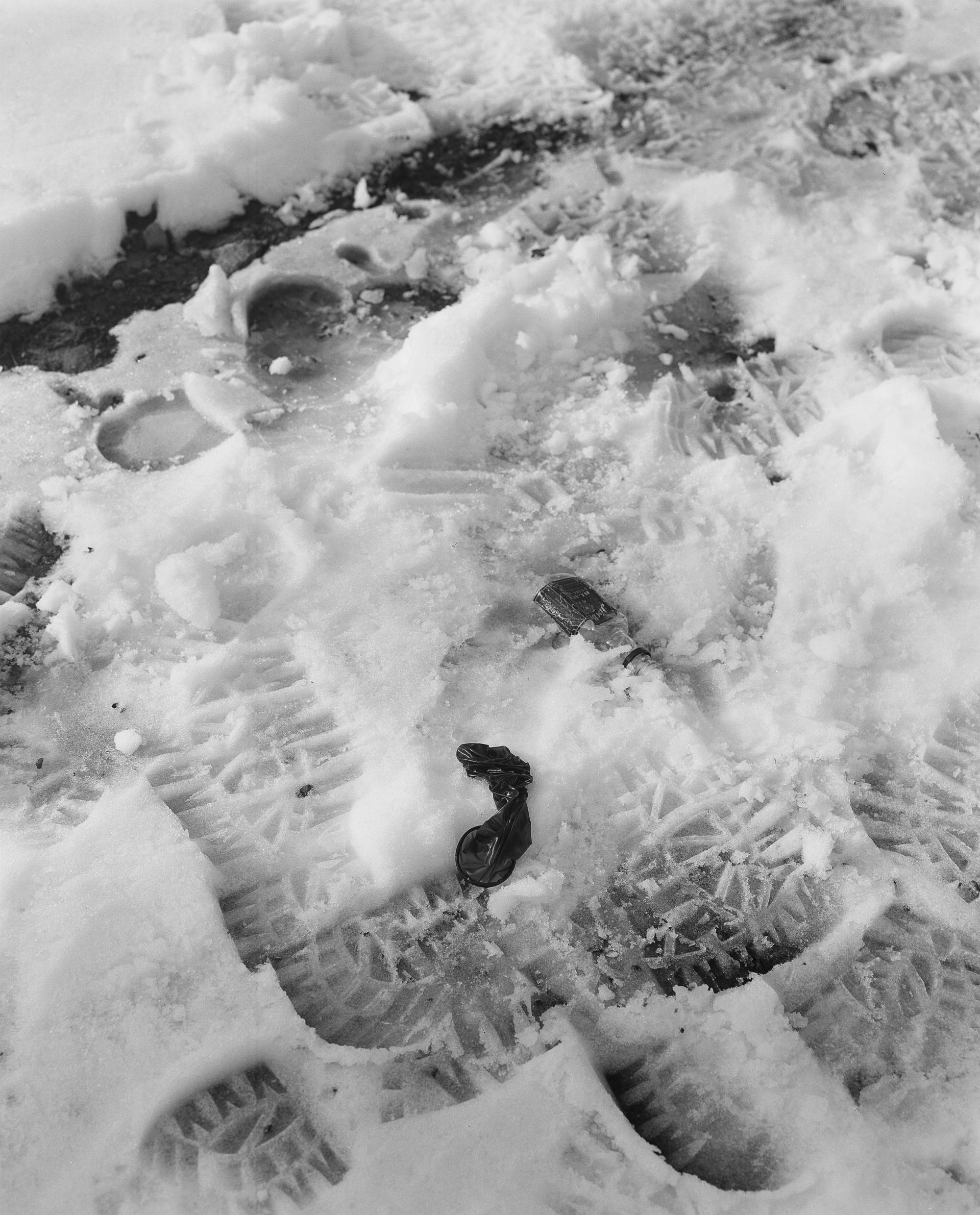 a black and white vertical image showing a used condom on top of foot prints in the snow 