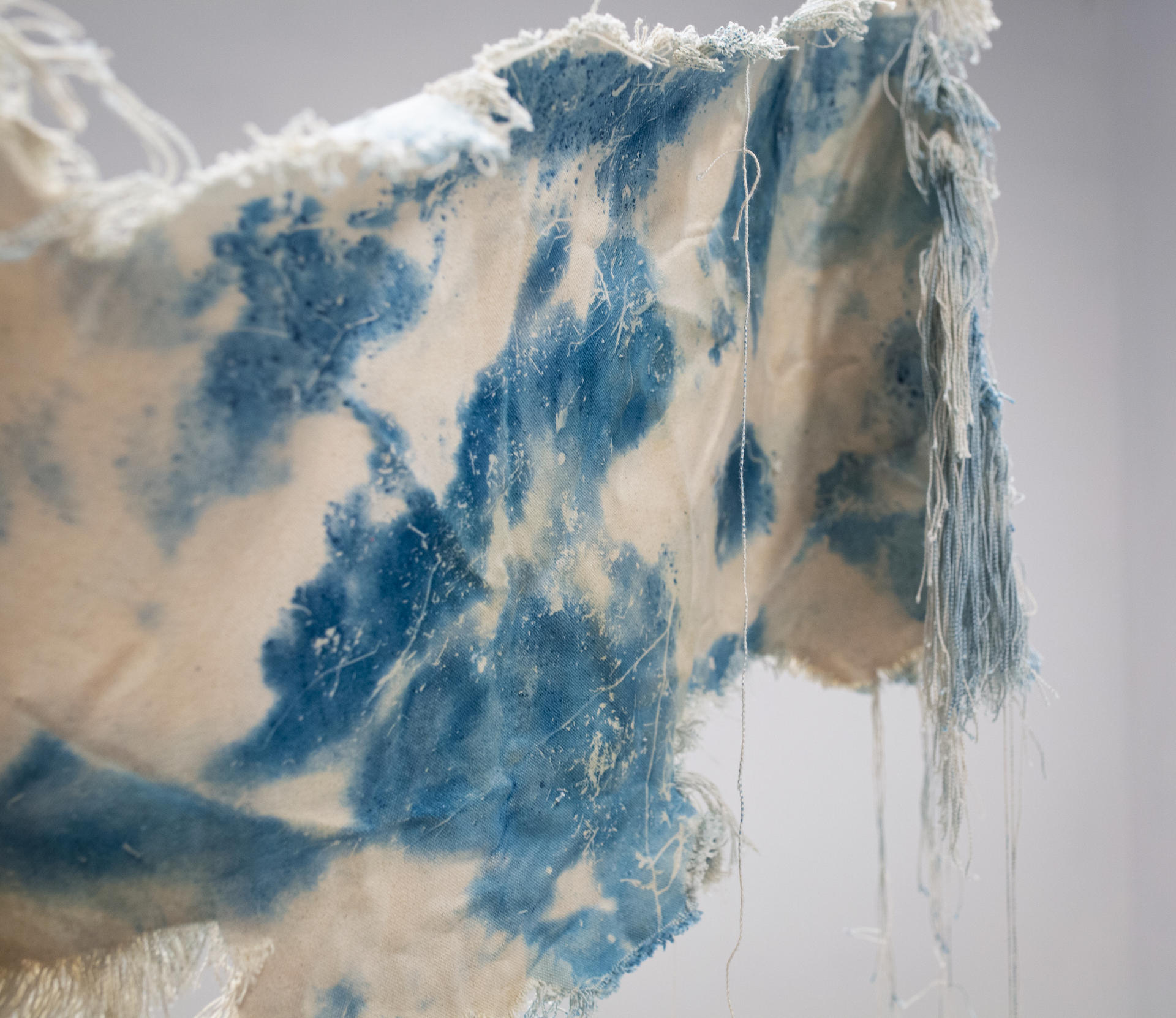 A beige canvas, dyed with spots of cyanotype is twisted and suspended in the air. The edges of the canvas have been unraveled and hang downward. 