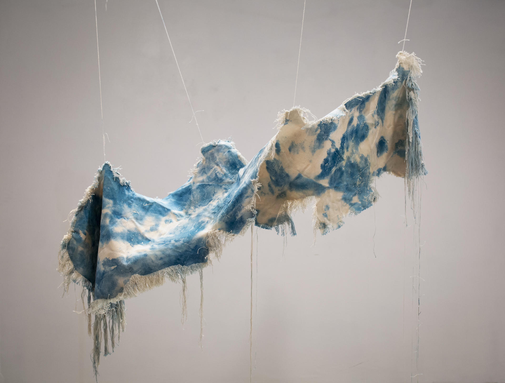 A beige canvas, dyed with spots of cyanotype is twisted and suspended in the air. The edges of the canvas have been unraveled and hand downward. 