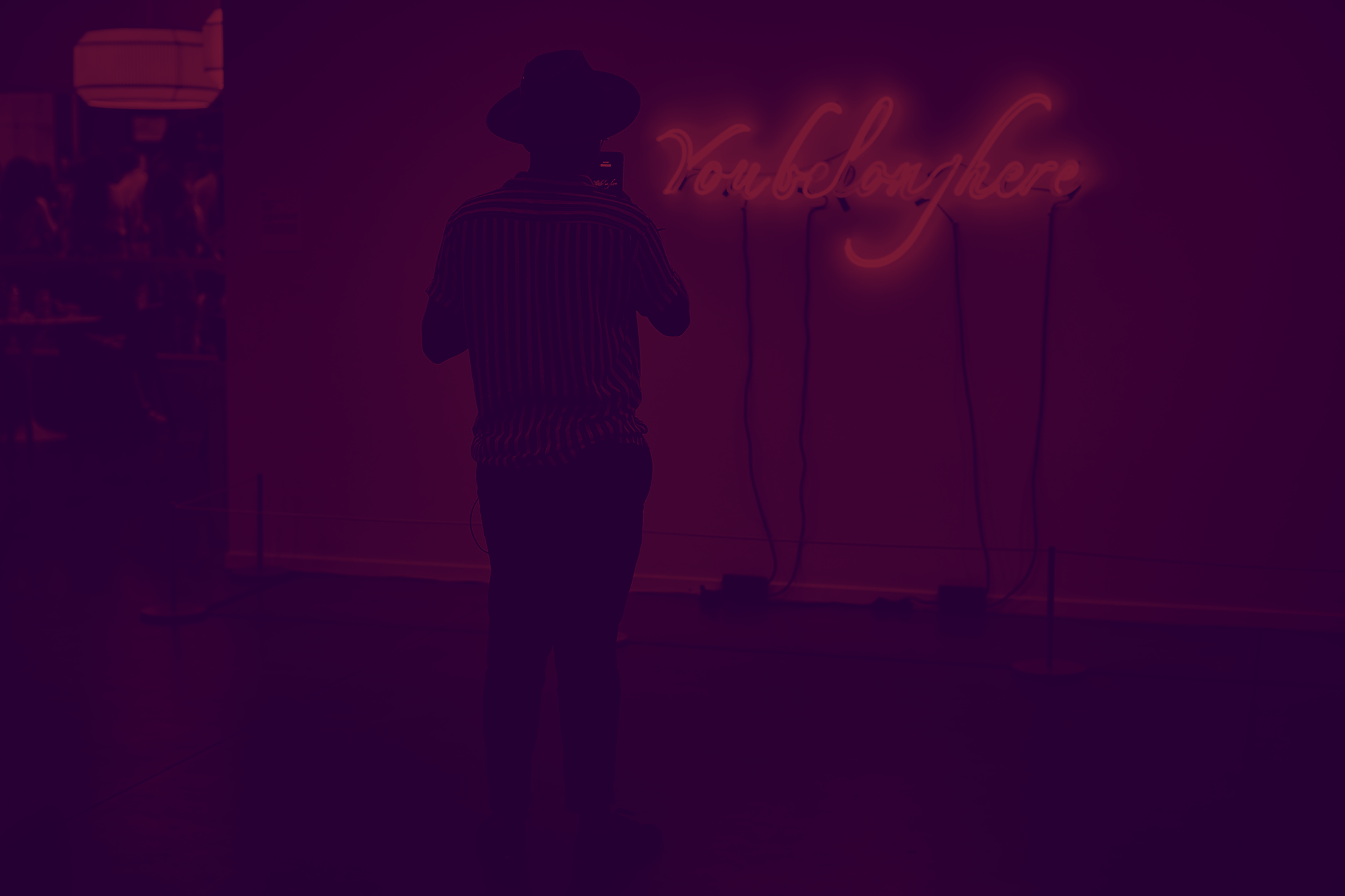 Standing with back to the viewer and in shadow, a person photographs a neon sign. It says, in light blue script, “You belong here.” A purple filter has been applied to the photo.
