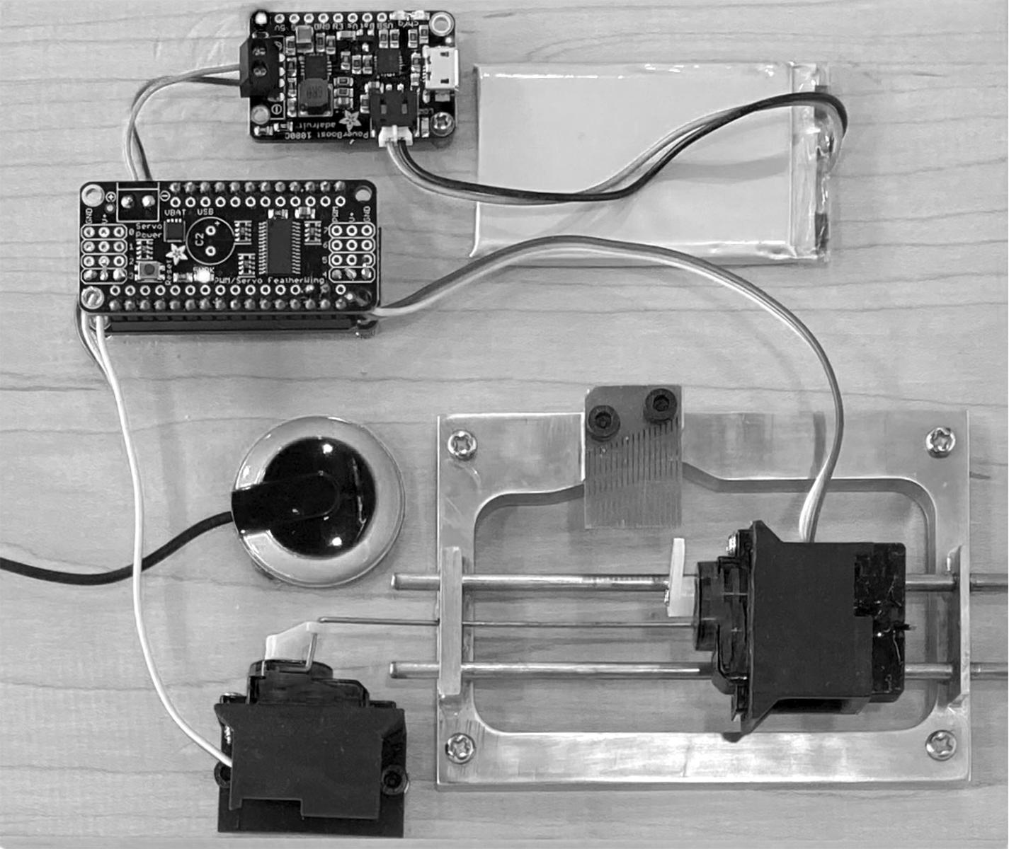 Image of a robotic music box. Two small circuit boards, a flat, rectangular battery, a round microphone, and a small motor are laid out around a metal frame which holds the music box tines and a motor on a track.