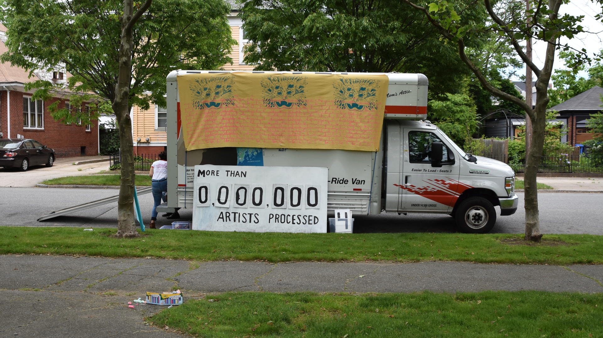 A U-haul van is parked by a park and some houses. A large banner with a brick pattern and three four-eyed lions hangs from the truck, and a big sign that says 'More that 0,000,000 artists processed' with replaceable numbers leans against the truck.