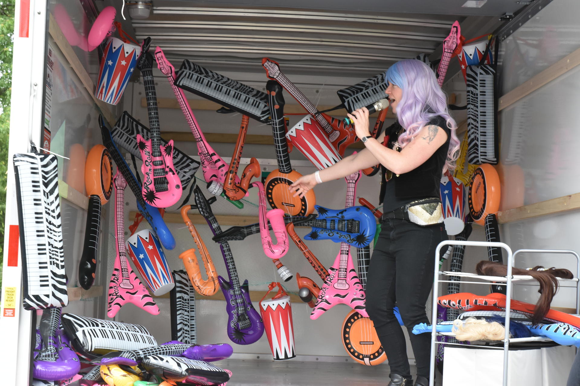 A white woman in a long light purple wig and a sequined fanny pack sings into a microphone while dancing in a U-haul filled with bright inflatable rock instruments