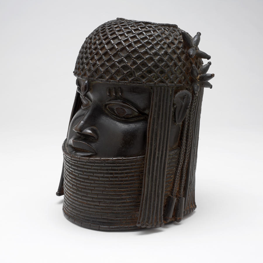 Elegant stylized portrait head. The subject wears an elaborately patterned headcovering and a tall stack of necklaces. Color is dark brown.
