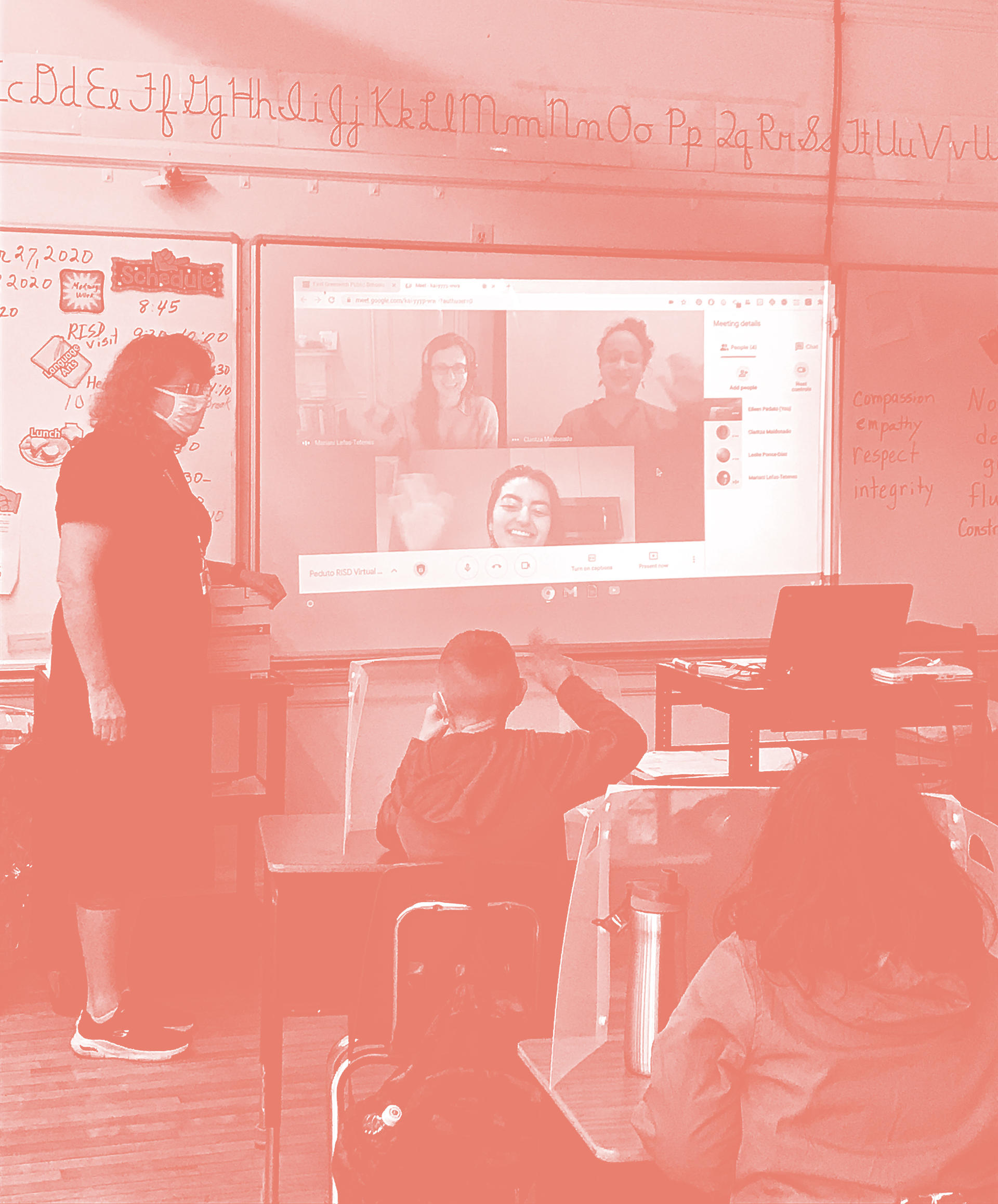 All-red photo of a classroom. A masked teacher stands beside a screen showing people on a Zoom meeting. A child in the front row of the class raises his hand. 