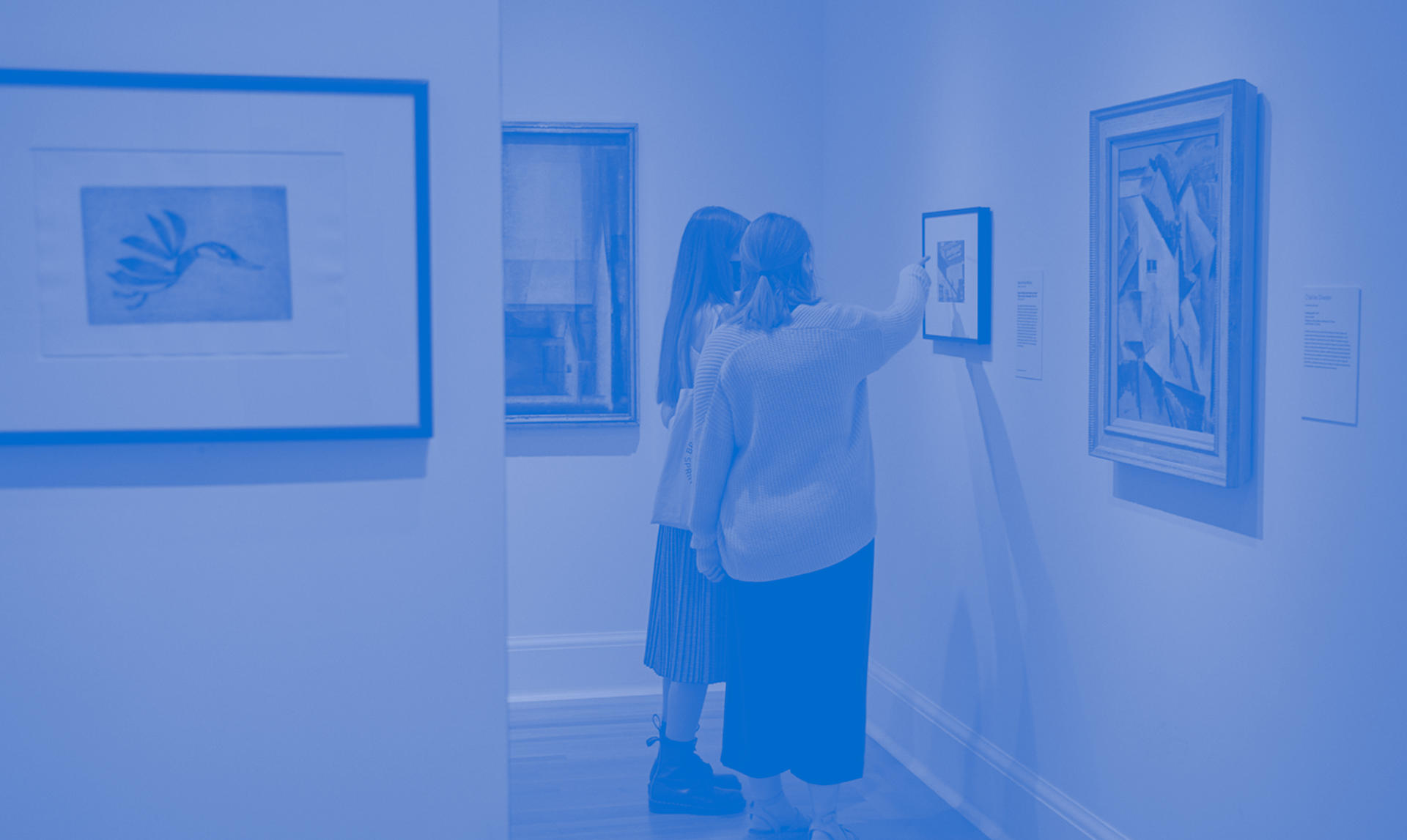 Blue-toned photo of two visitors in a gallery space. They stand in front of a small framed work. The figure on the right gestures to it.