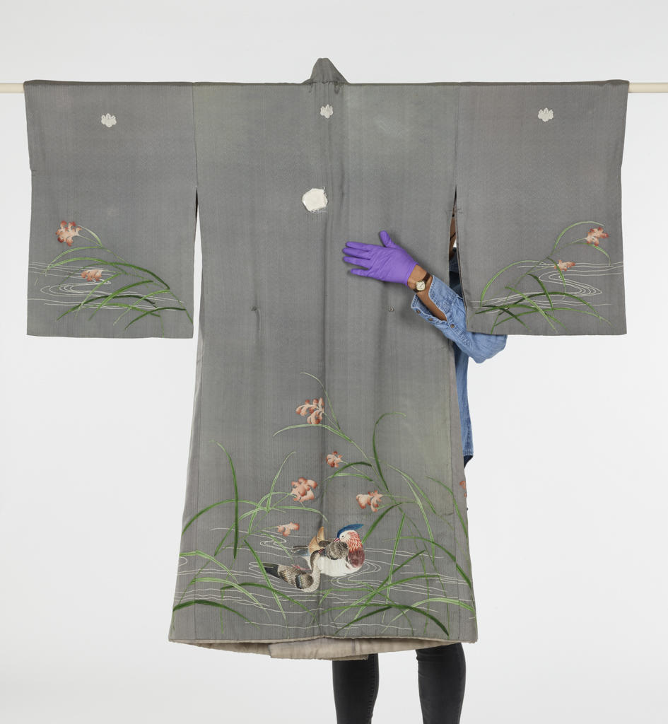 An embroidered gray Asian robe hangs from a rod. A museum worker stands behind it, looping one purple-gloved hand to the front. The result is something like an embrace.