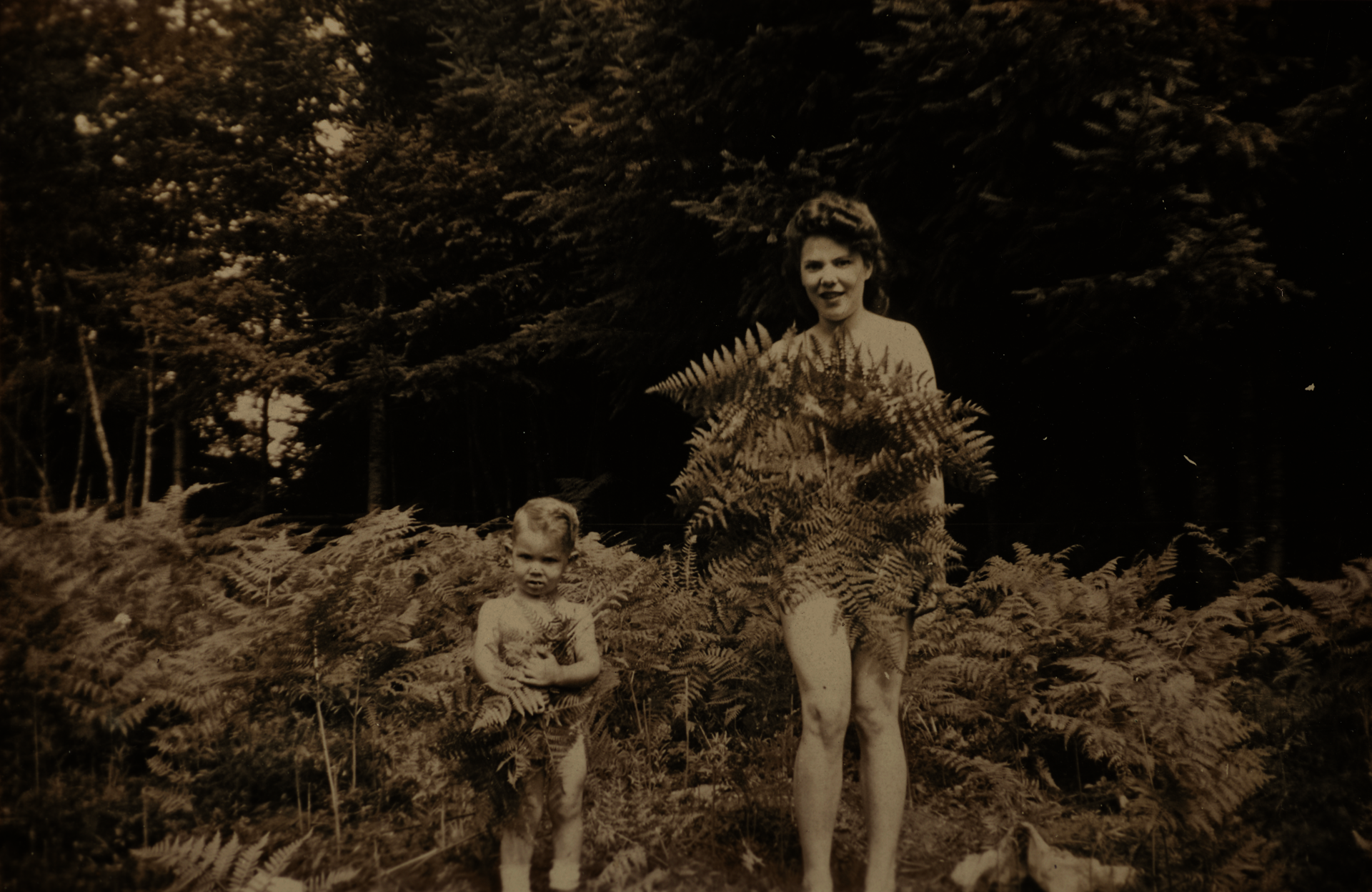 Snapshot of a light-skinned woman and young child standing naked in a bed of ferns at the edge of a forest. The woman smiles, both covering themselves with fern fronds.