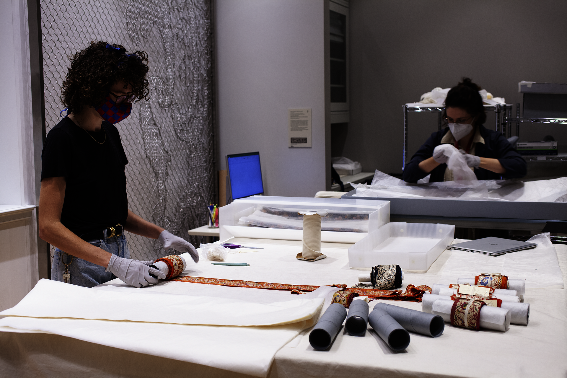 A masked museum worker stands at a table, materials laid out before them. A second person works sitting at a table in the background