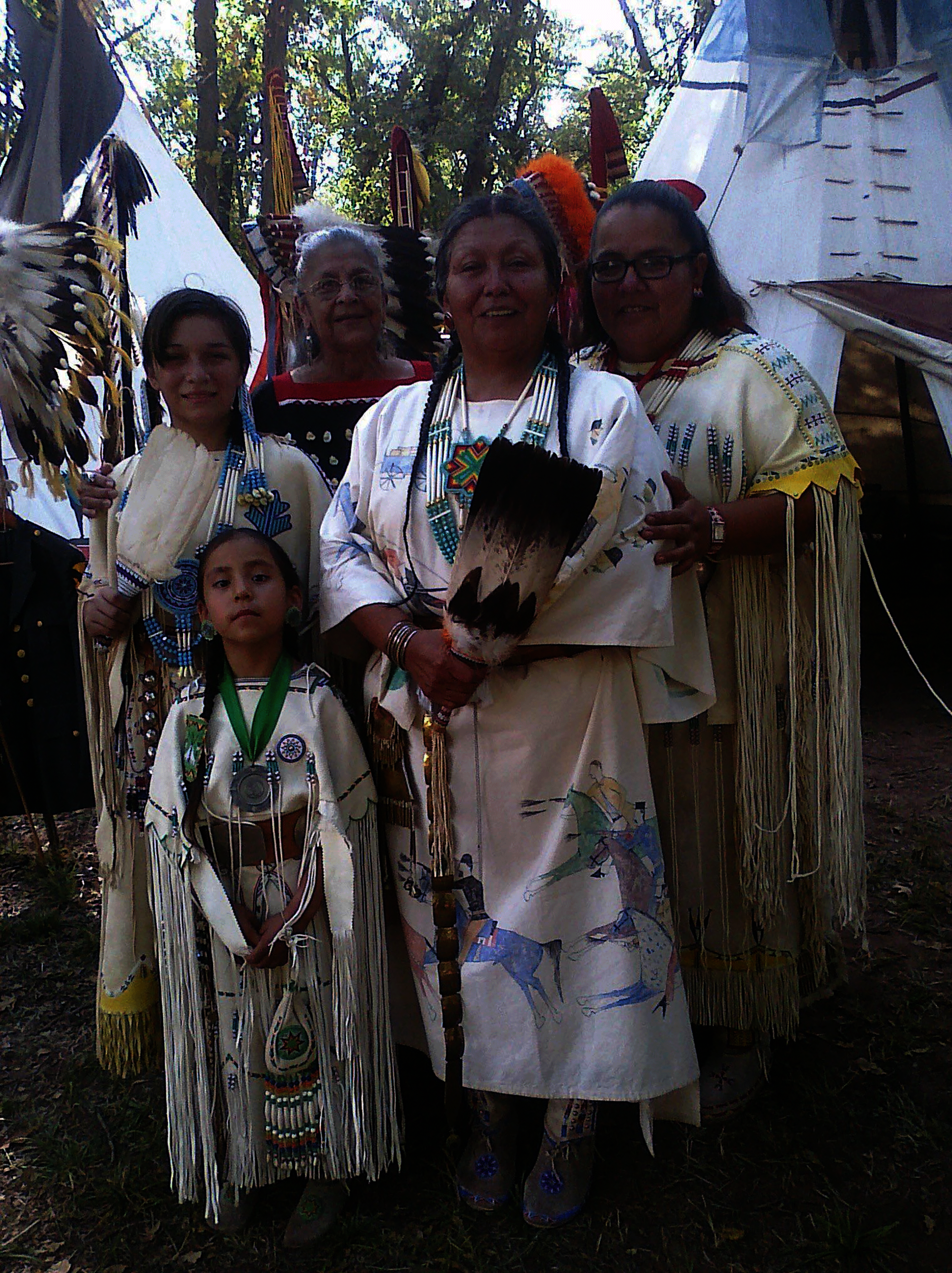 Five Indigenous women and girls stand between two tepees. They wear decorated buckskin dresses and hold feather fans. They look at the viewer, and several of them smile.