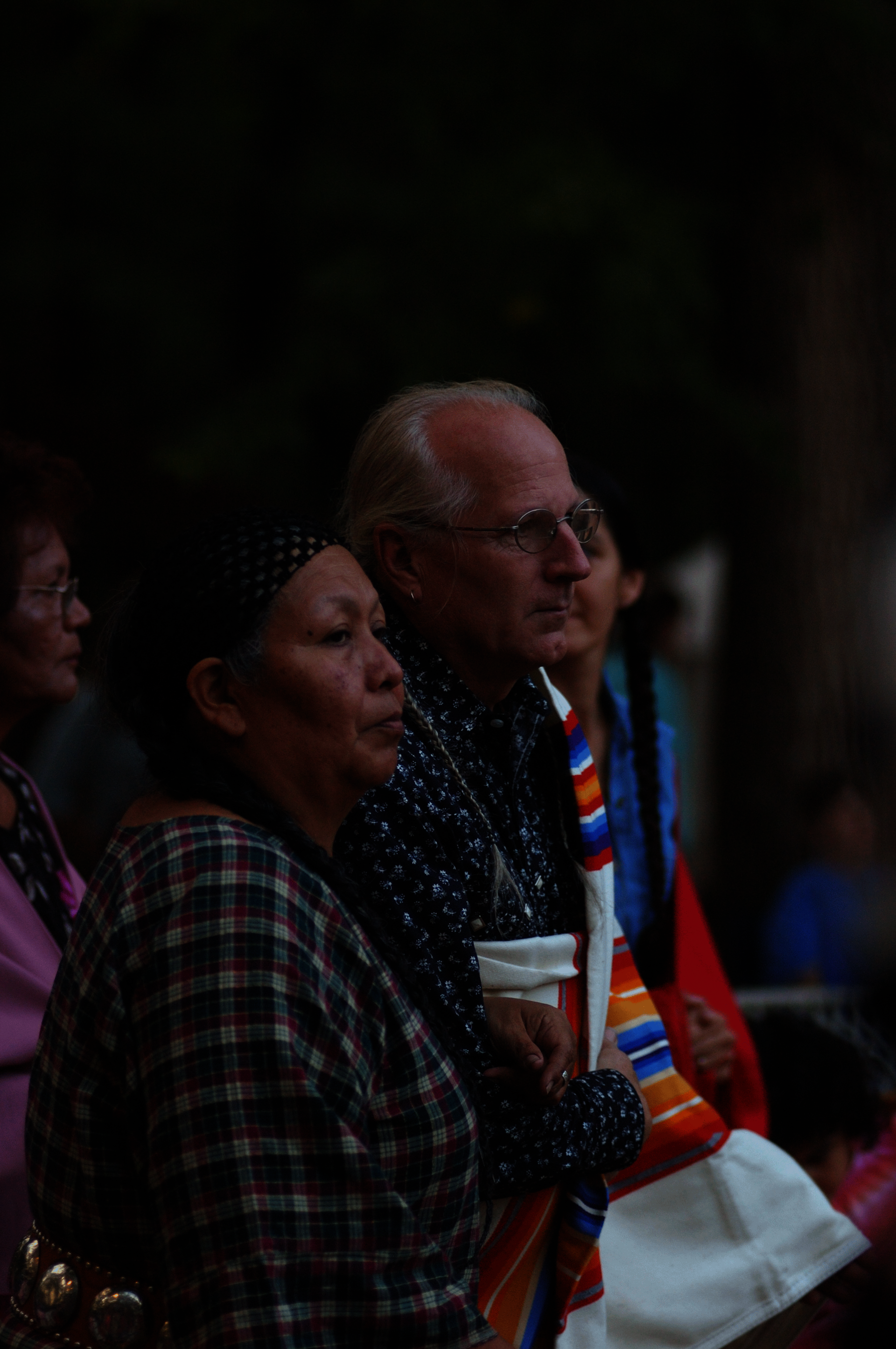 Photo of an older couple in profile. She wears long dark braids, a bandana, and a plaid shirt. He has glasses and white hair and wears a colorful striped textile over his shoulder.