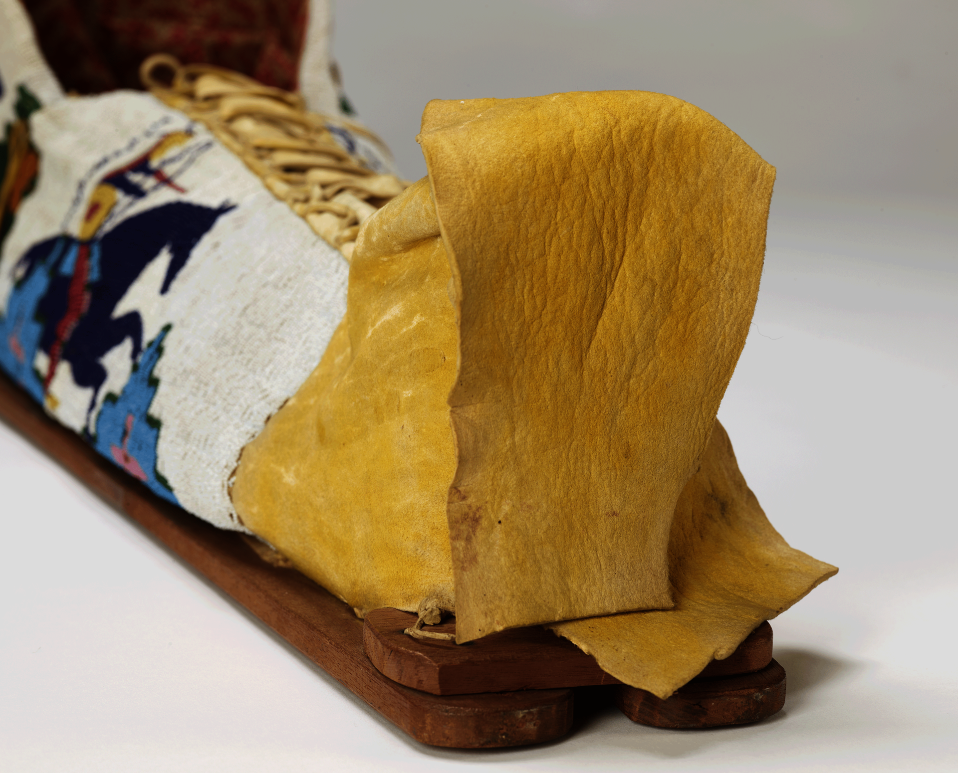 Detail of beaded object with wood supports at bottom. A yellow leather panel is loosely folded, attached to a panel densely beaded with blue horse and rider. Top is laced closed.