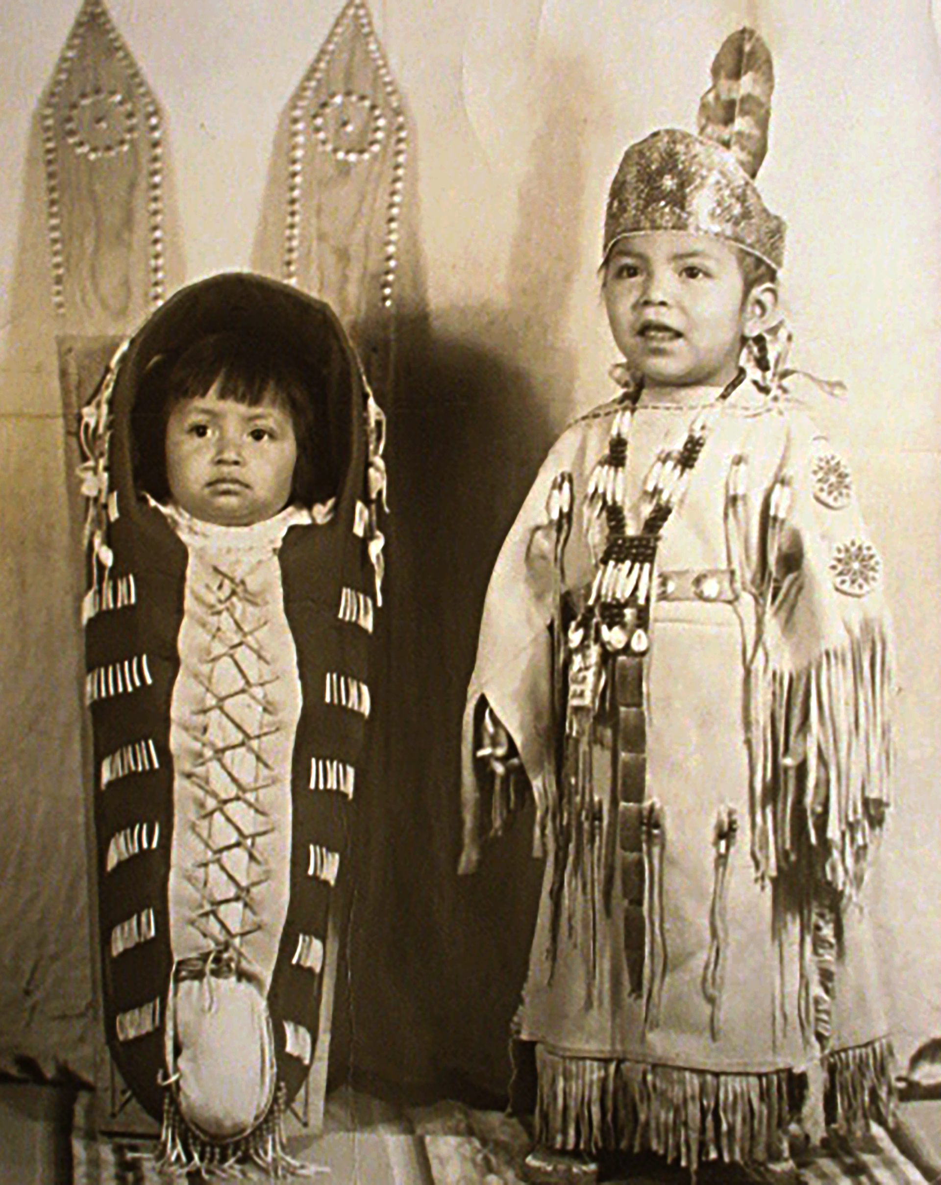 Black and white photo of two Indigenous children. A baby is laced into a carrier at left, and a toddler in a beaded buckskin dress stands at right.