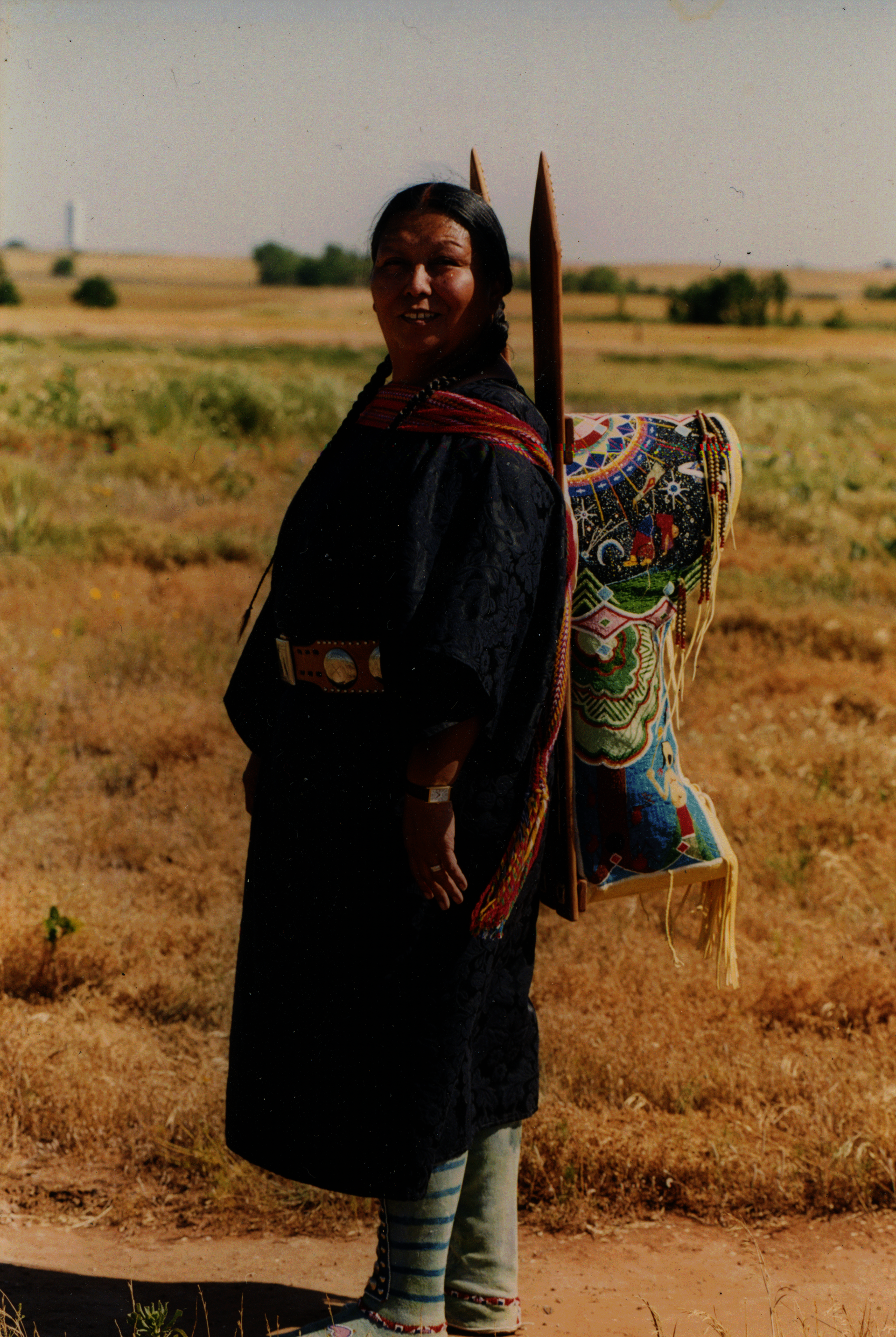 Brown-skinned woman wearing a colorful baby carrier strapped to her back. Her hair is braided and she wears a dark dress and striped socks.