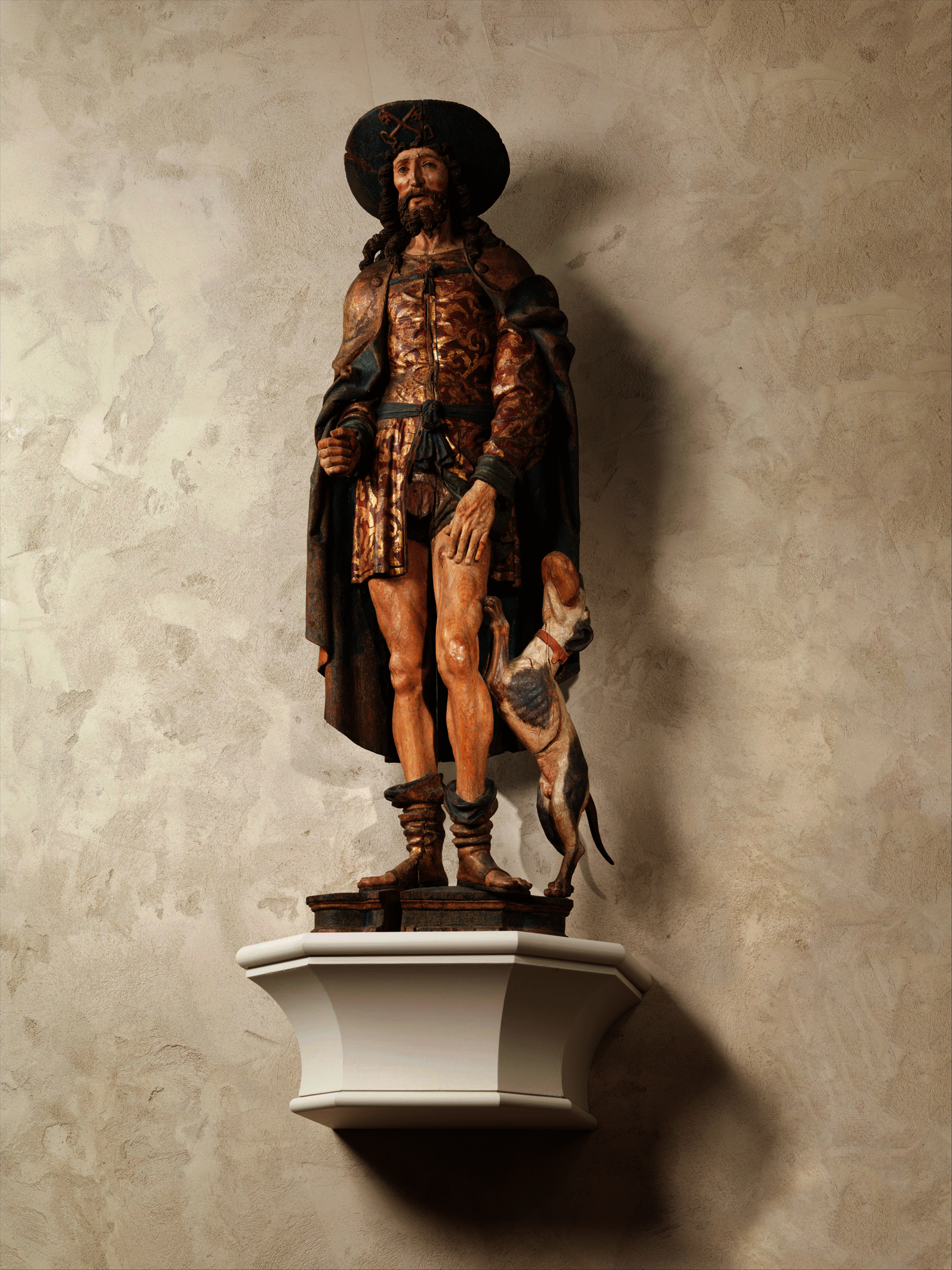 Painted sculpture of a bearded man in an elaborate short tunic and cloak. His left hand rests on his bare thigh. A dog stands on his back legs beside him.