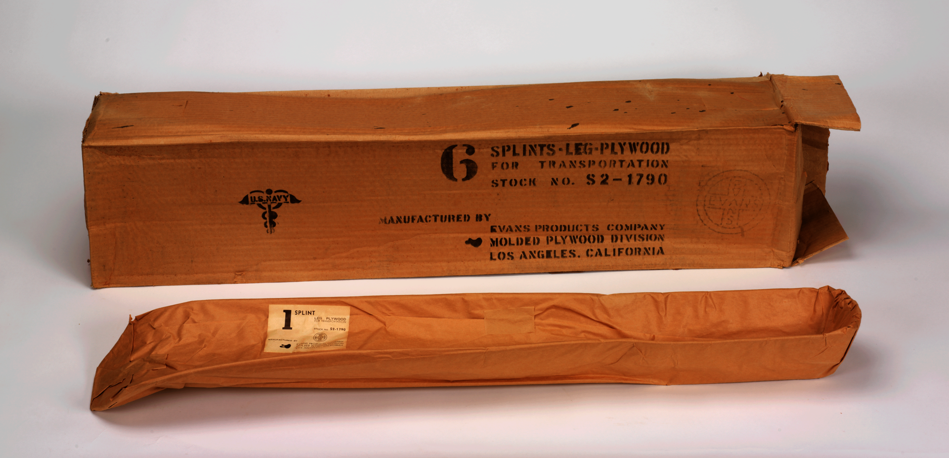 Photo of a wooden support made for leg injuries, wrapped in yellowed paper. Behind it is a cardboard box labeled with the details about the device.