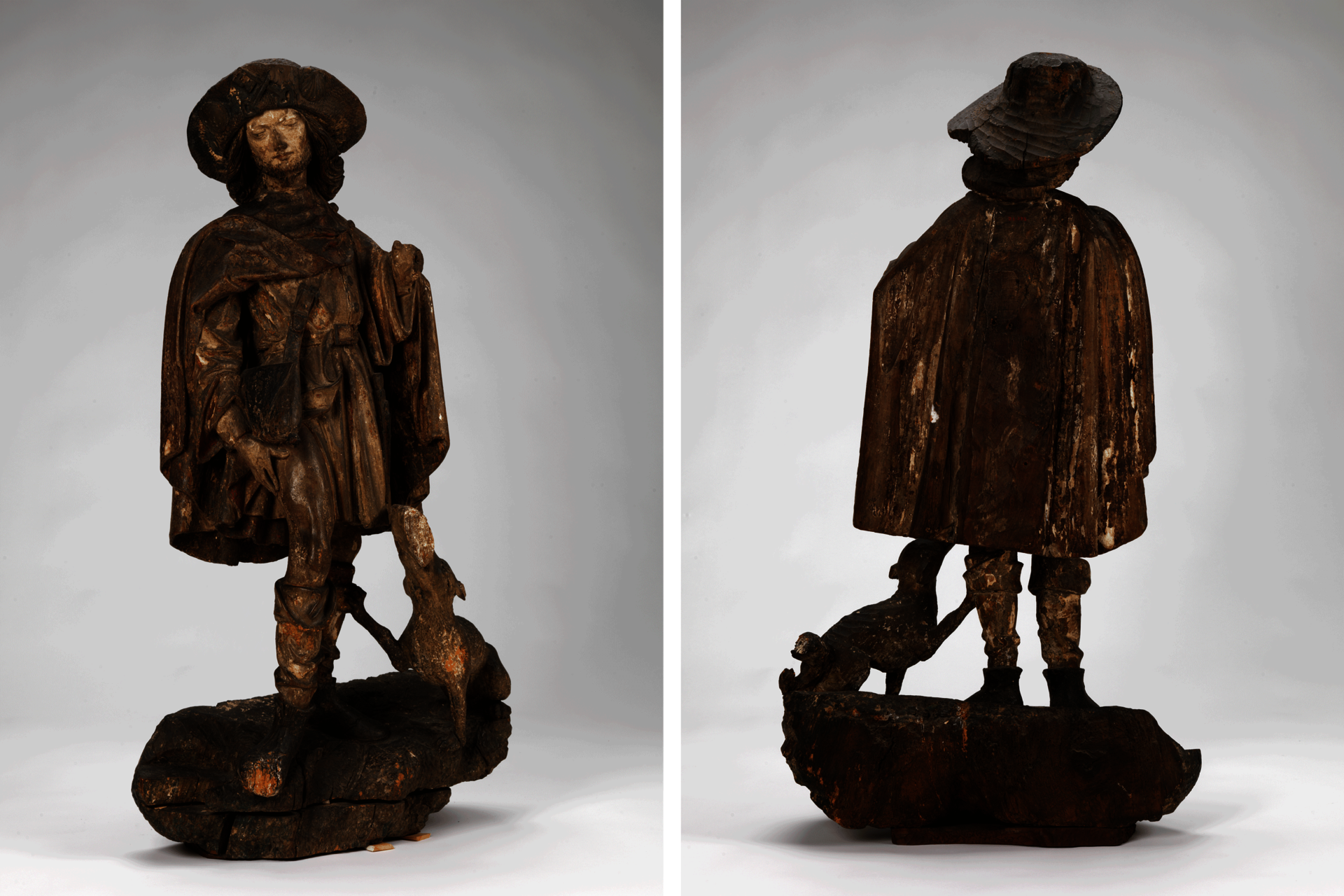 Dark-brown sculpture of a standing man wearing a cape, hat, and boots. He looks to the side, hand on his thigh. A dog at his feet looks up at him.