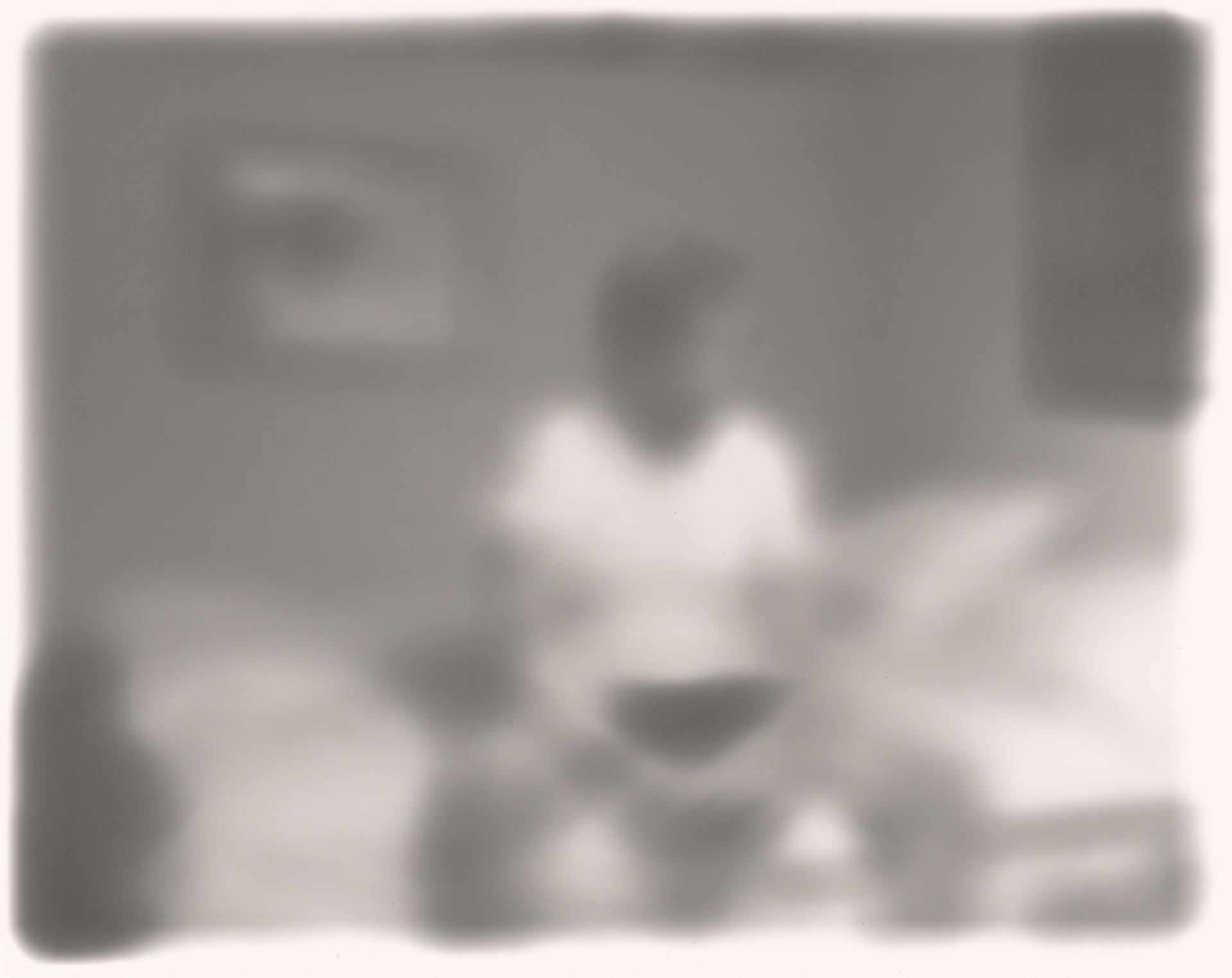 Dreamy, out-of-focus black-and-white image of a figure sitting on a bed, hands clasped between their knees.