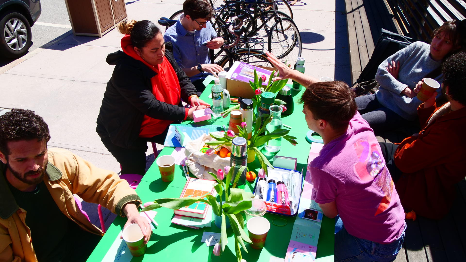 6 students gather around a green table covered in flowers, drinks, paint, and a books of drawings and photographs.  