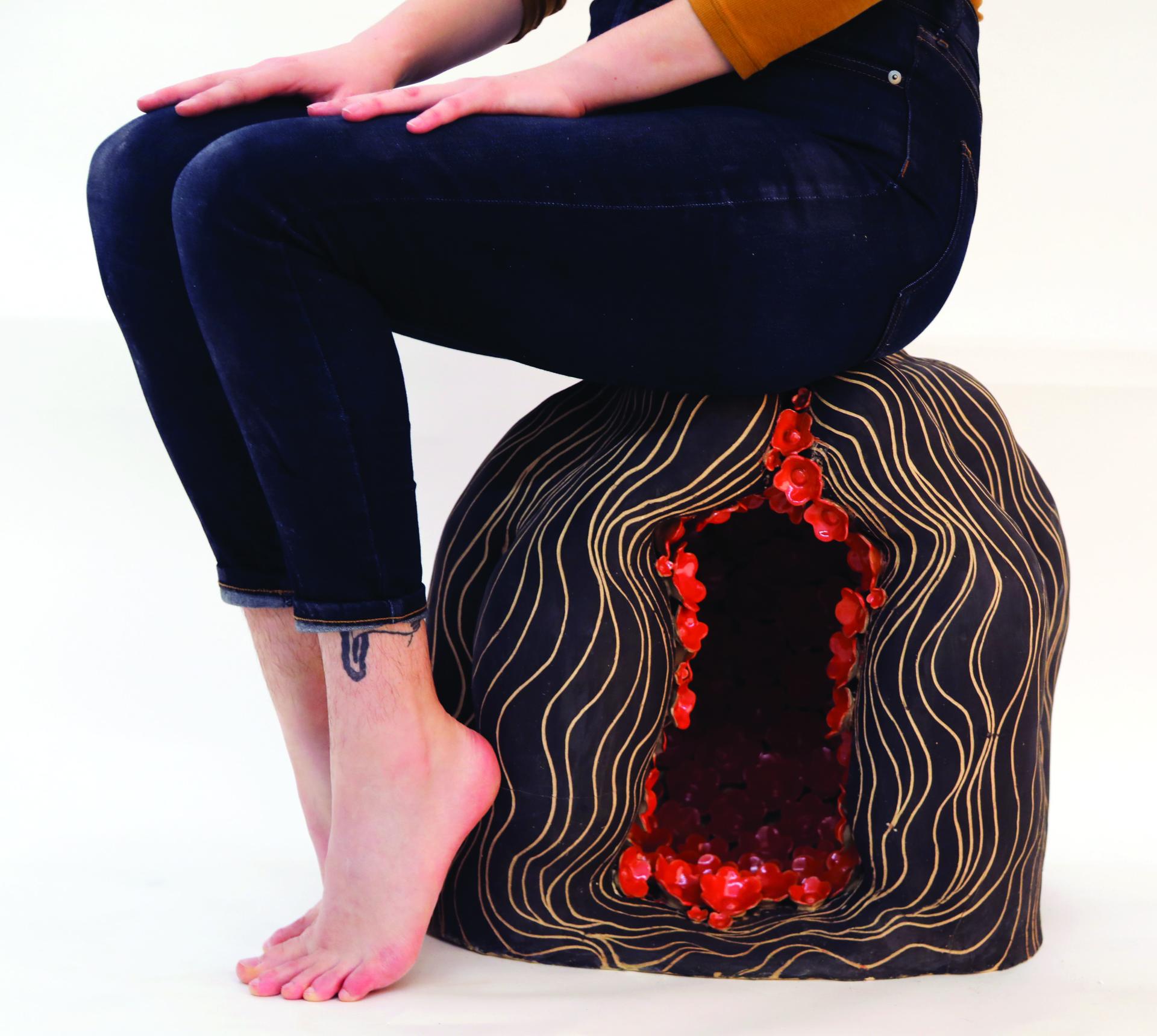 Image of a person from the waist down sitting on a ceramic head with red flowers inside its cavity.