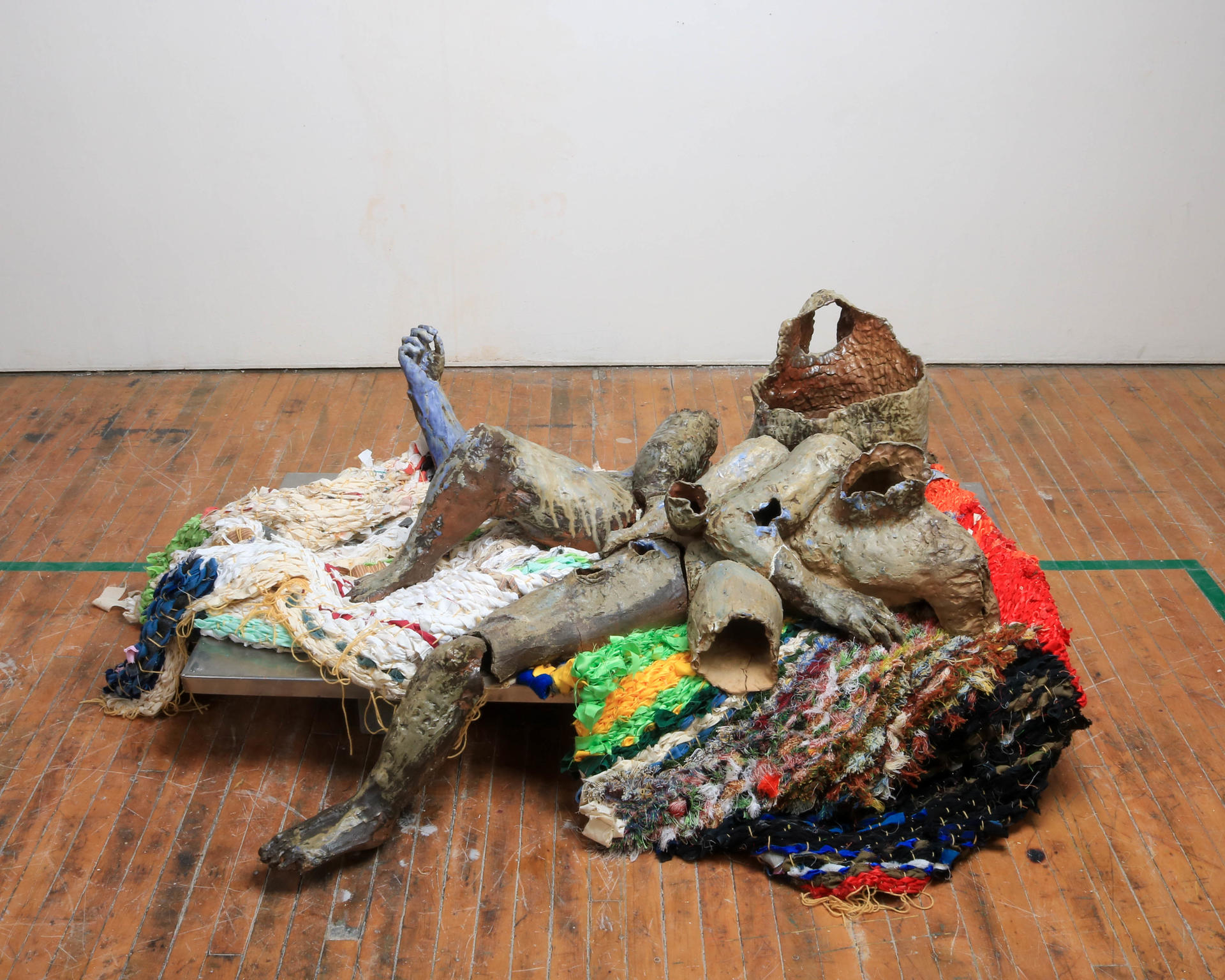 A multi-limbed, fragmented ceramic body reclined on two colorful woven fabrics atop a steel plane. 