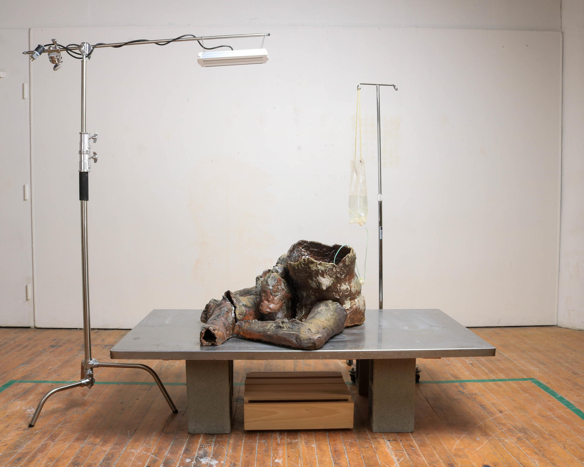 A dark, abject ceramic body with only two abstracted legs sits upon a steel slab with an IV Bag and grow light above.