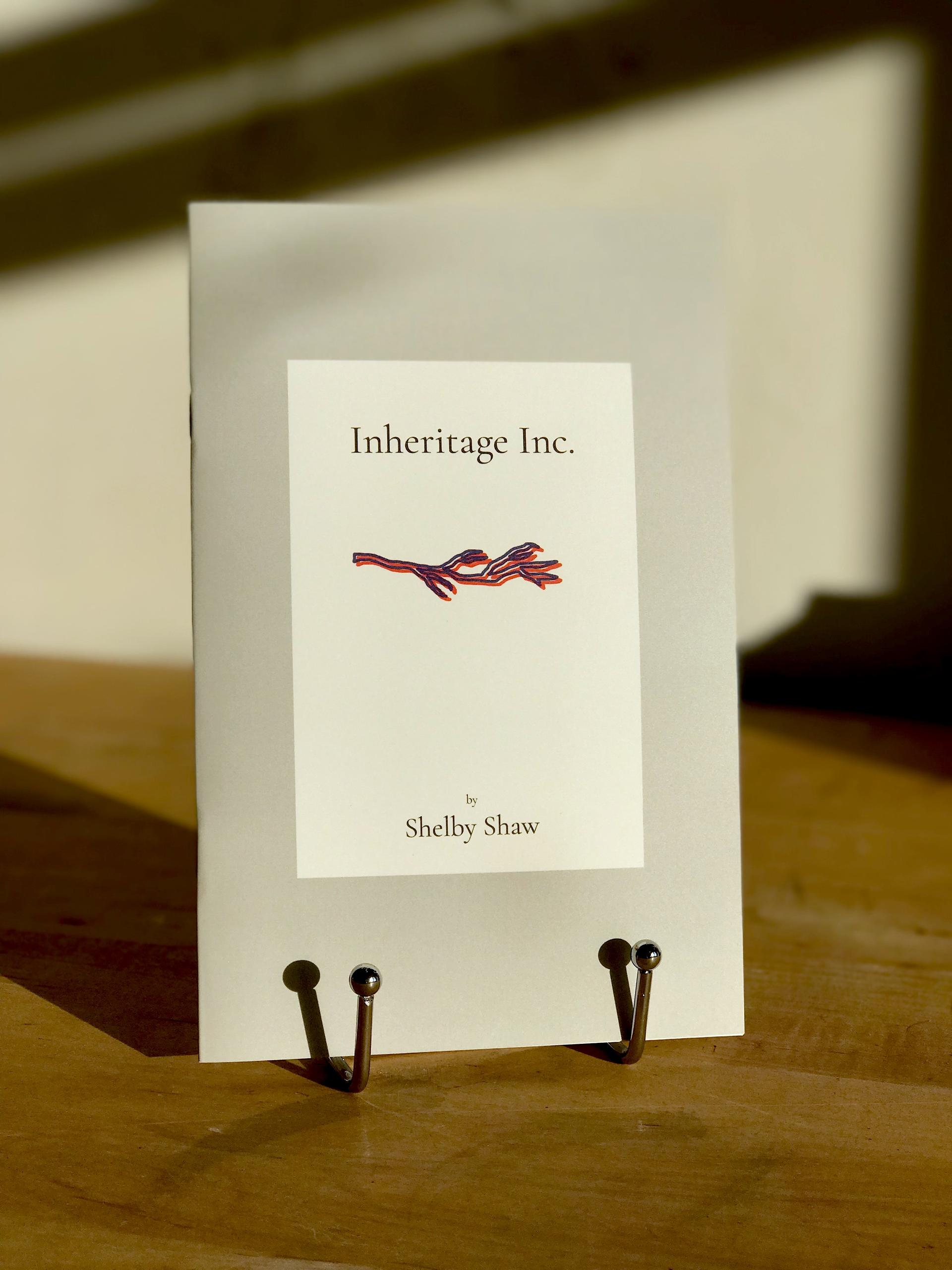 The book 'Inheritage Inc.' which is a grey color with white center rectangle containing the title in black font and a red and blue illustration of a tree branch, is displayed on a silver book stand atop a wooden tabletop with a white wall in the background.