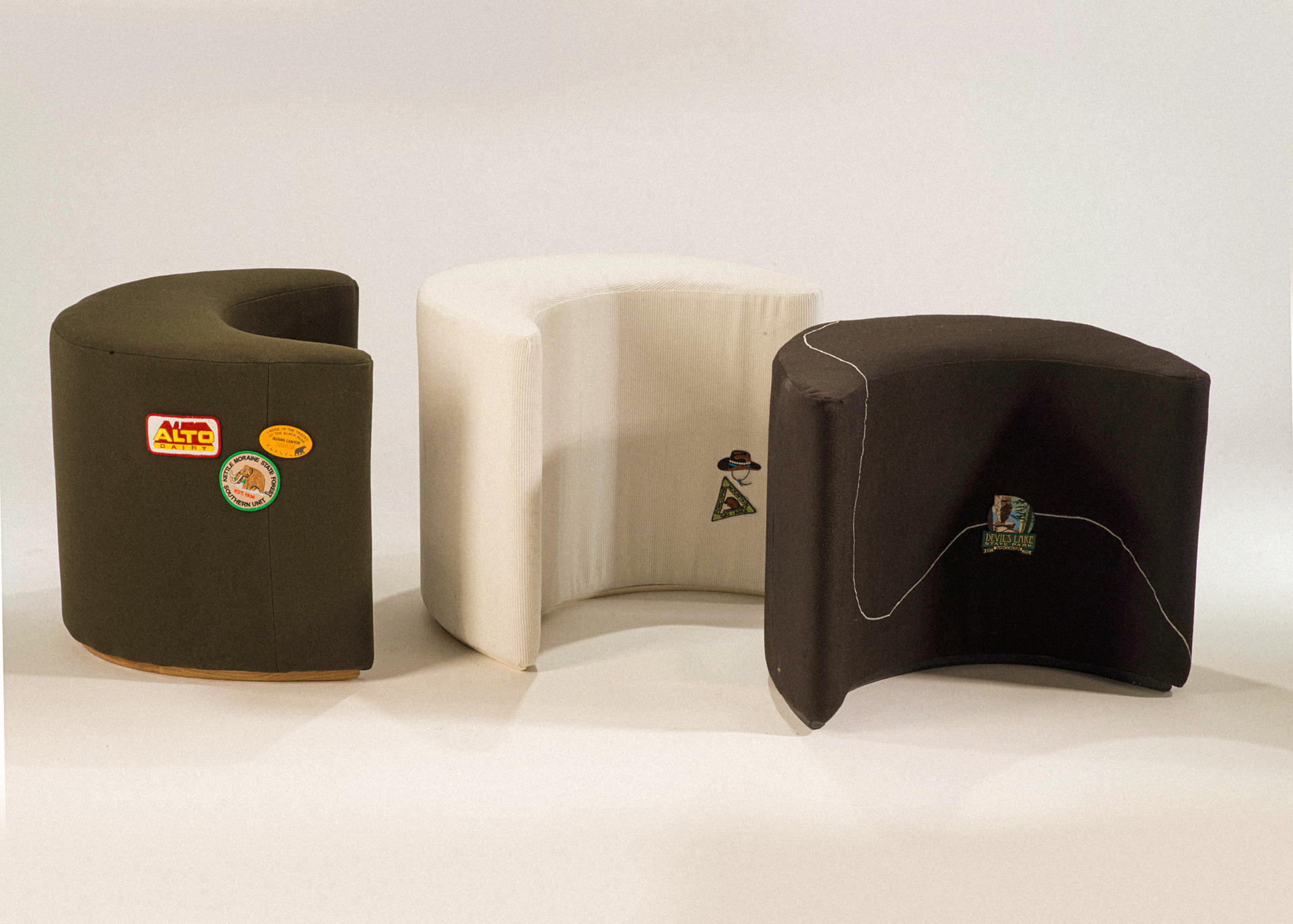 three crescent-moon shaped upholstered stools: one black, one deep green, and one white corduroy. embroidered patches are applied to the surface of all three.