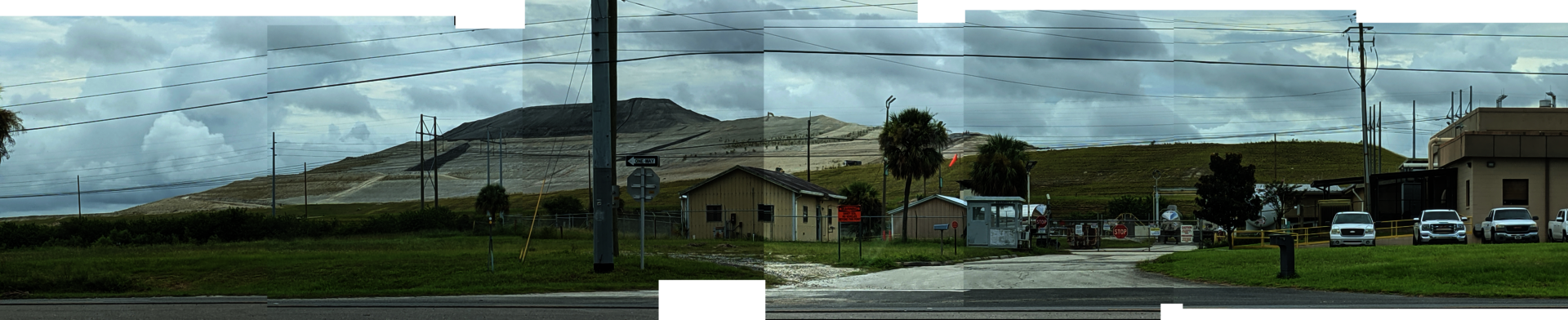 A massive mound of grey phosphogypsum rises in the backround against a cloudy sky. In the foreground, some buildings and vehicles serve to convey the scale of towering gypstack. 