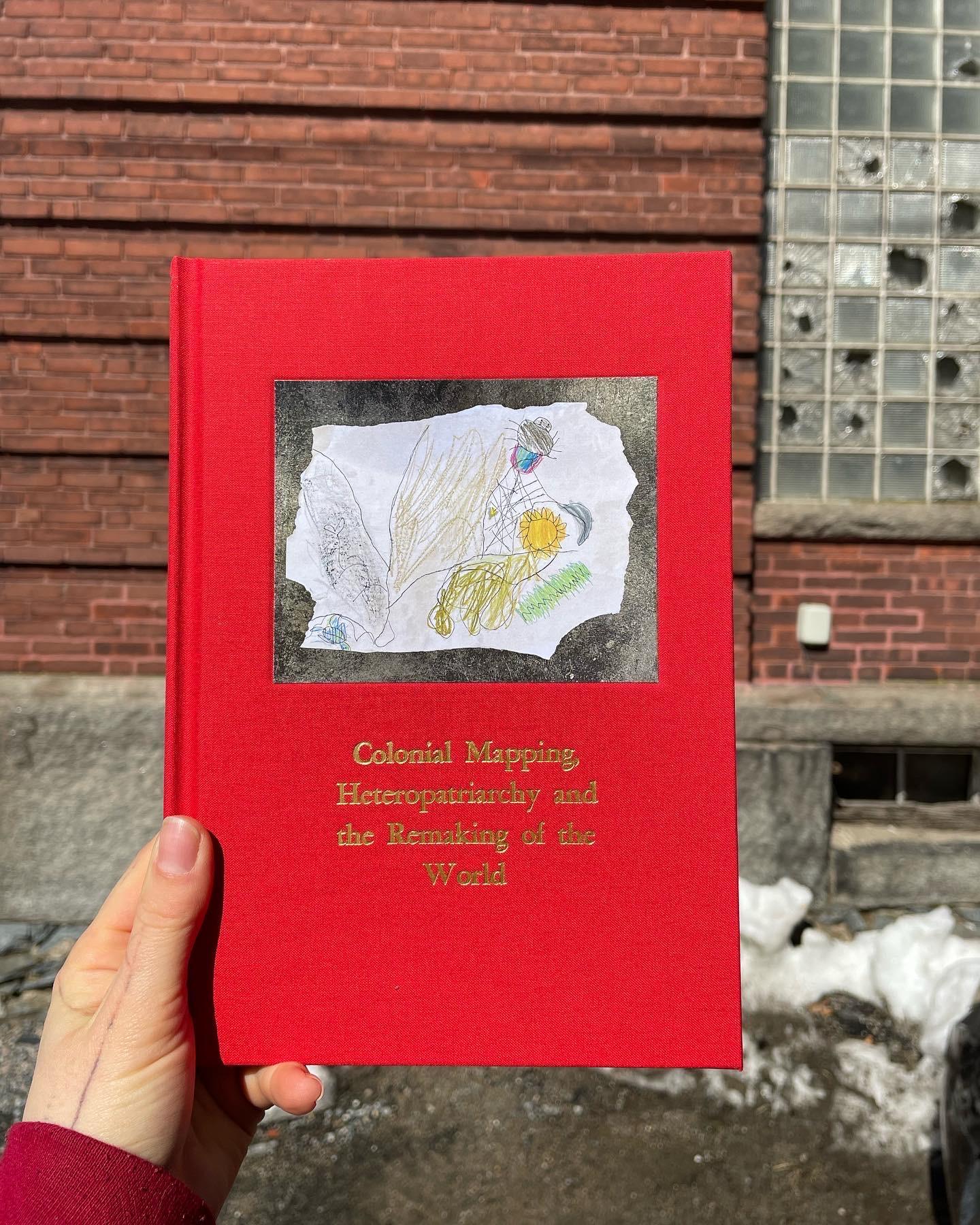 A hand holding a red book titled, "Colonial Mapping, Heteropatriarchy, and the Remaking of the World", with an image of a drawing.