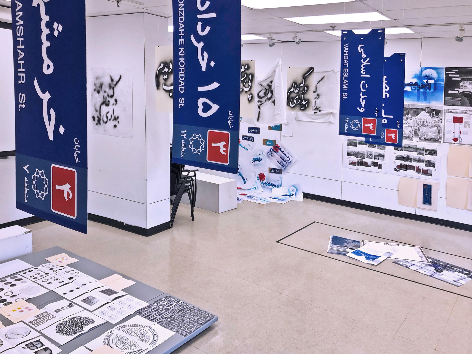 This image shows a multi-component installation in a room with objects hanging from the ceiling, printed outs pinned on the wall, and books laid on the ground 