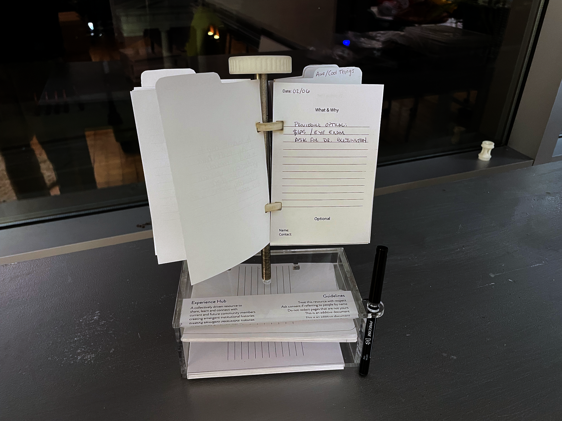 A small steel pole holding half sheets of paper that people have written on. The pole is on top of an clear box holding extra paper and a pen. 