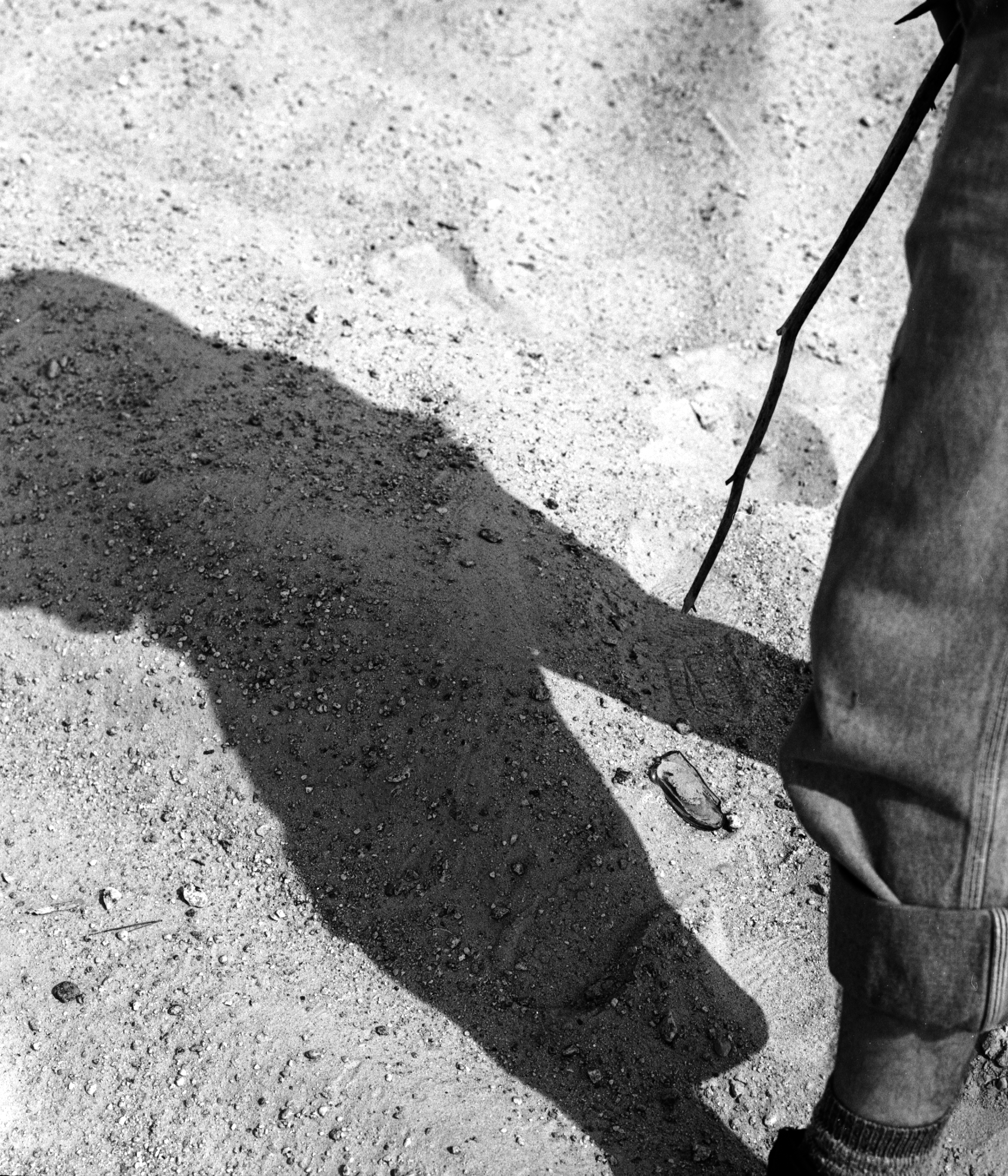 A black and white photograph of a piece of glass in the sand between two legs with someone pointing a stick from above. 