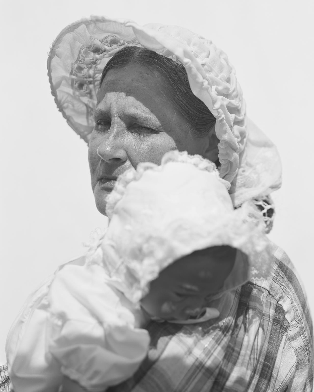 A woman in 19th century clothes stands against a white backdrop in the bright sun. She looks at the camera with one open eye. She is carrying a baby in her arms. 