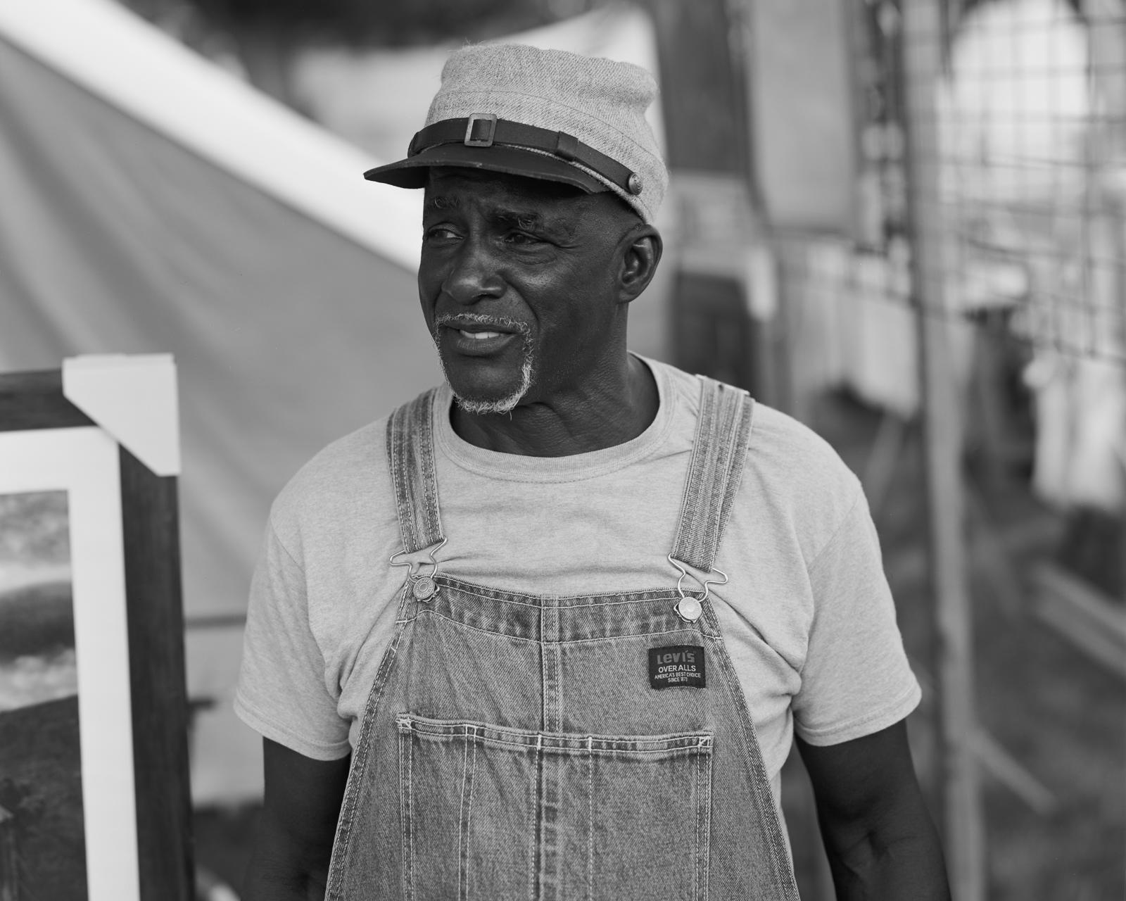 A Black man stands in the center of the frame looking off to the left, he is wearing a Confederate hat. Behind him are canvases and frames and posters. 