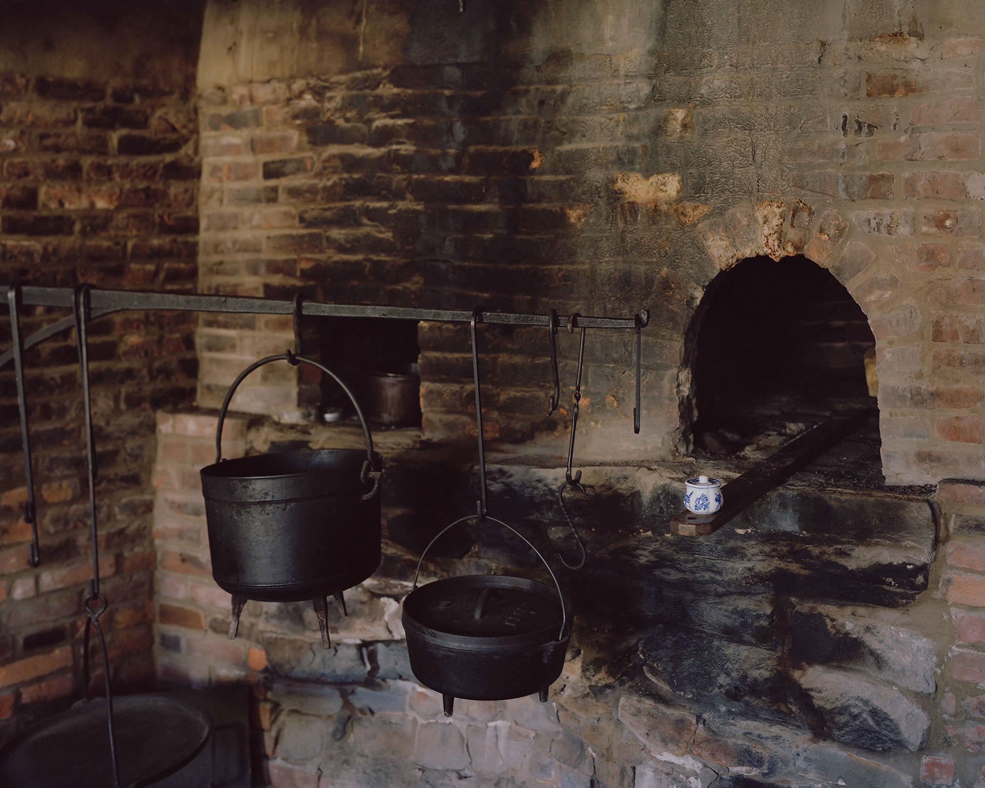 Photograph of brick oven hearth, with three cast iron pots hanging from a large rod. 