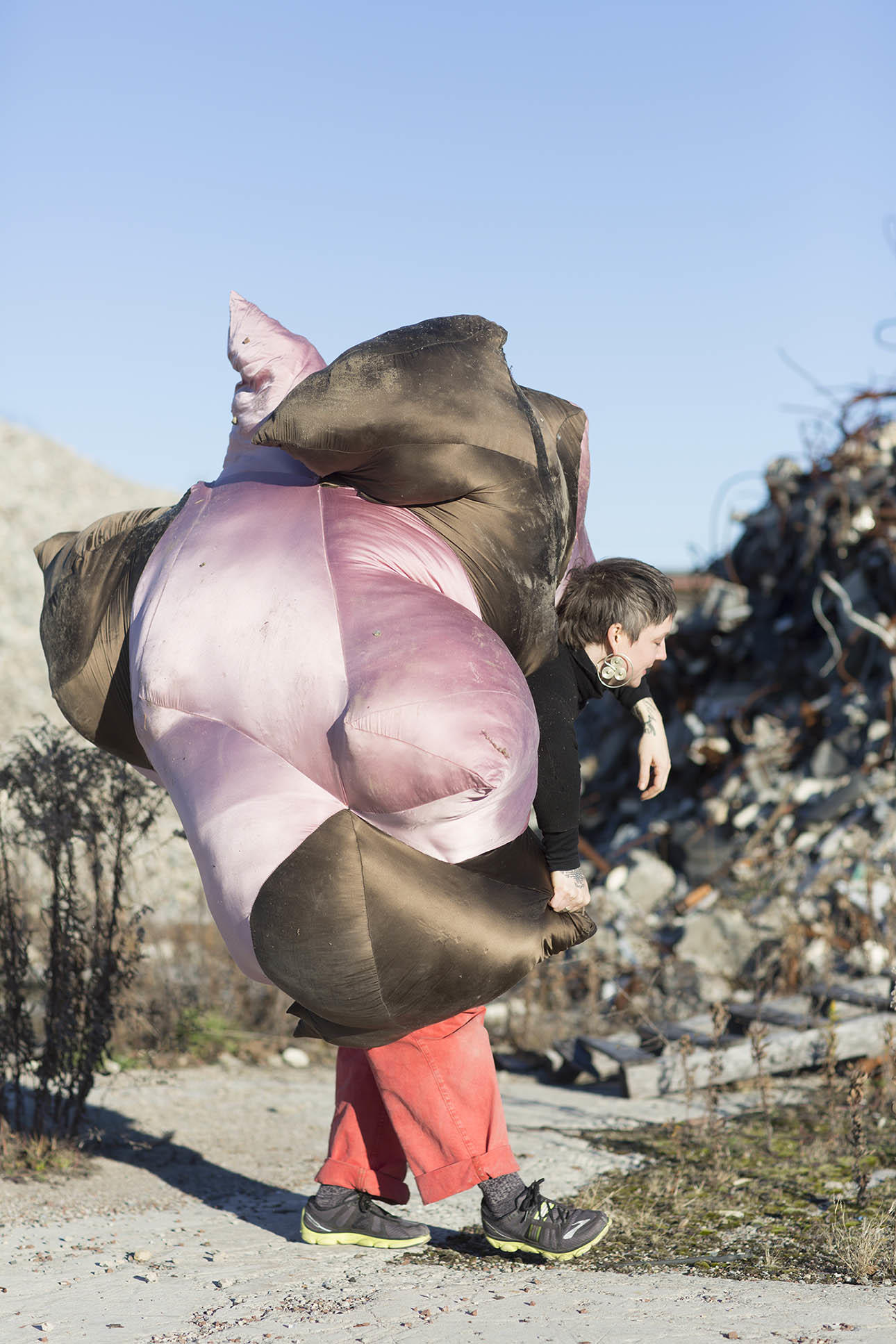 A person with short salt and pepper hair wearing red pants carries an immense and strangely shaped pillow-like object amongst pieces of tattered rebar and broken steel.