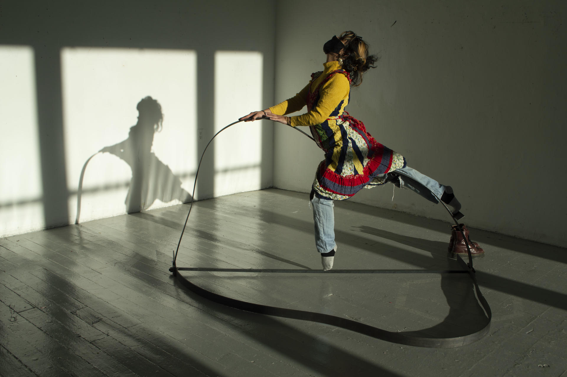 A person with dark hair and a colorful dress layered over jeans balances and dances atop a delicately bent steel beam.