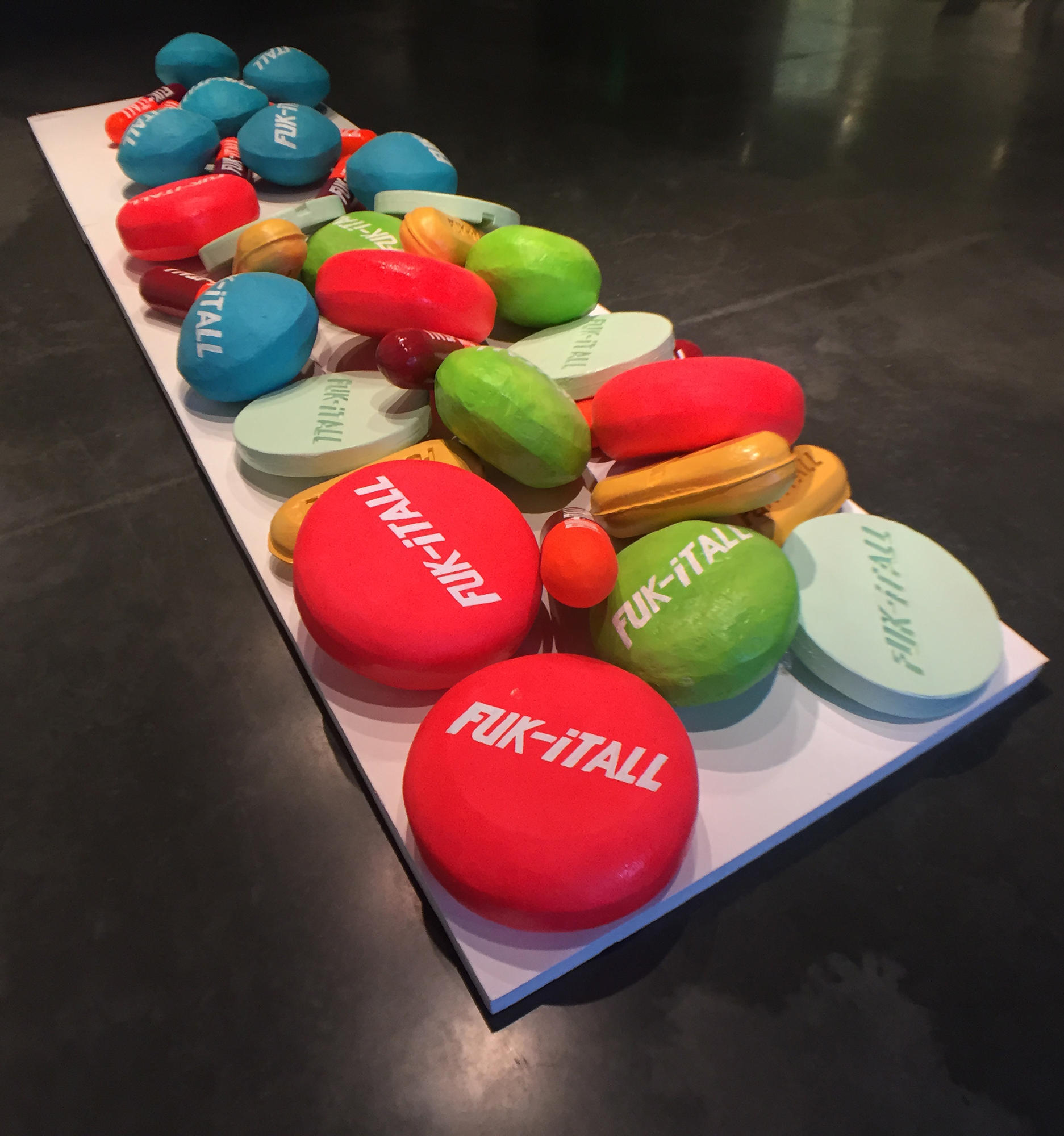 The work is an assortment of brightly colored basketball-sized pills which are of varying lengths, shapes, and designs. These pills are placed on a stretched-out platform that takes up a prominent area in the center of the floor. It is made of mixed media of rubber and wood on foam board.