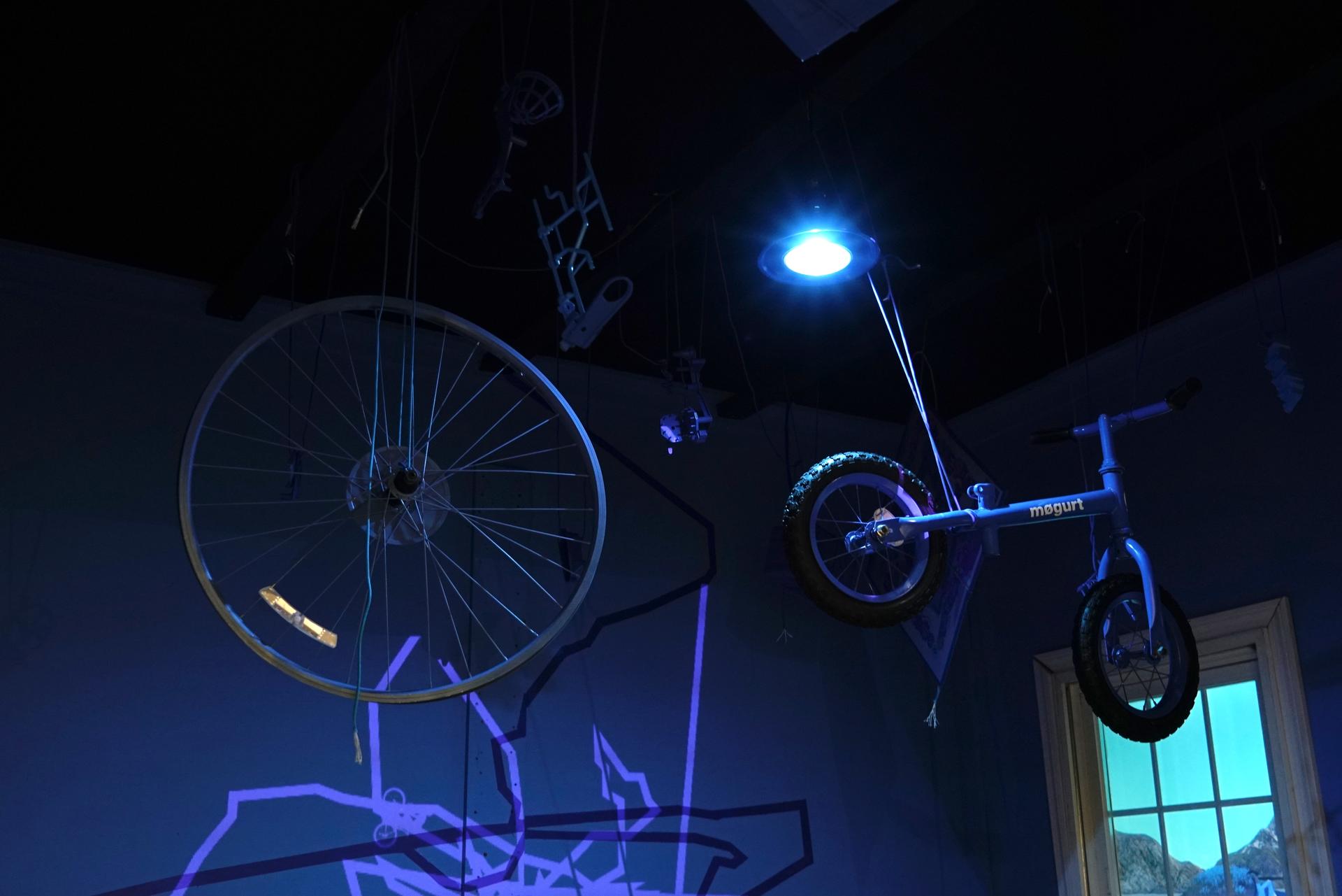 Installation materials suspended from the ceiling: Mouse trap board game pieces, kid's bike, busted bike wheel, paracord