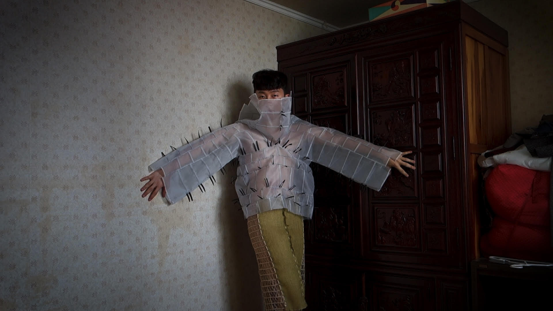 Model poses in a dimly lit corner wearing a shirt made of translucent white squares and yellow pants.