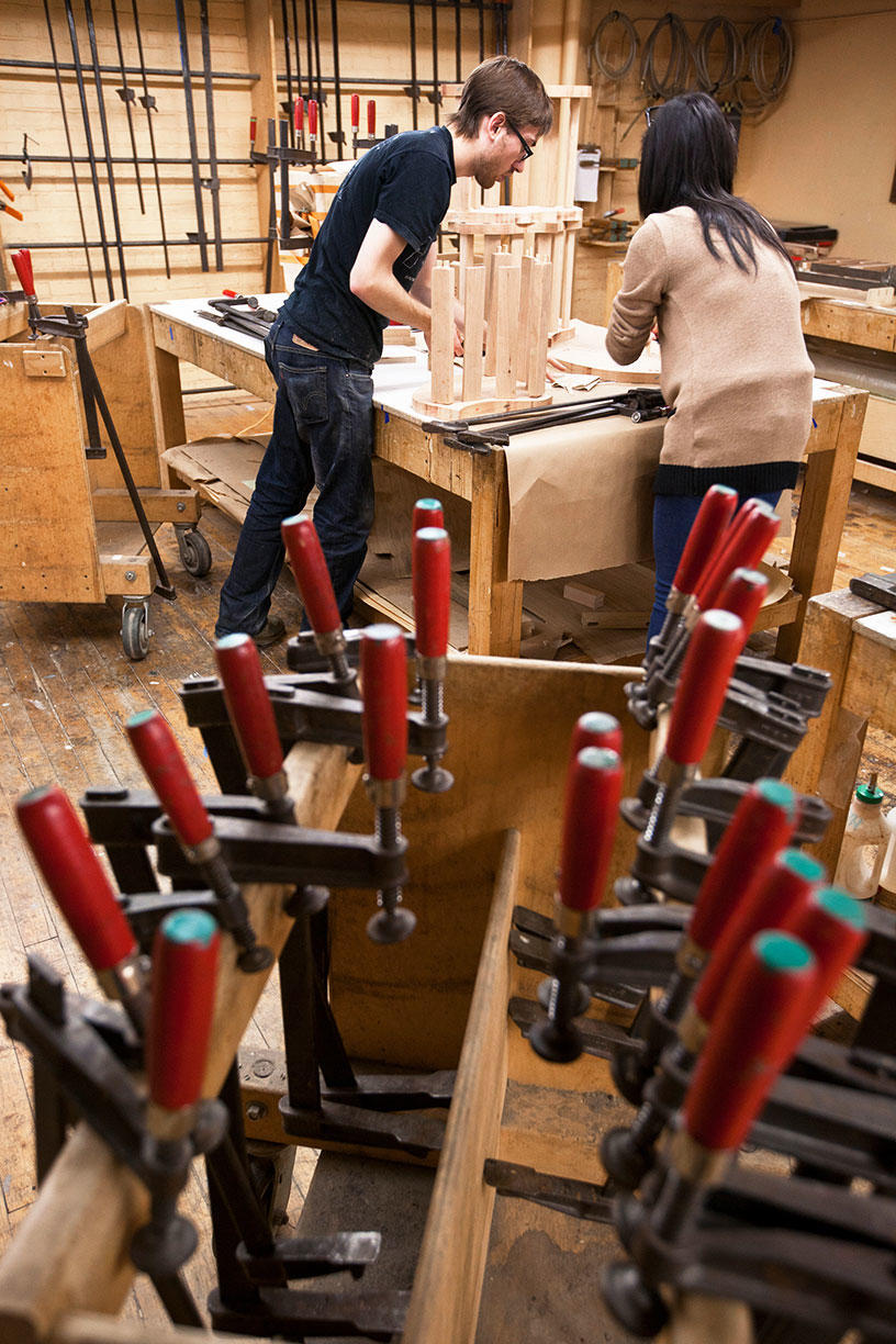 Two people working in a woodshop, with vice clamps in the foreground