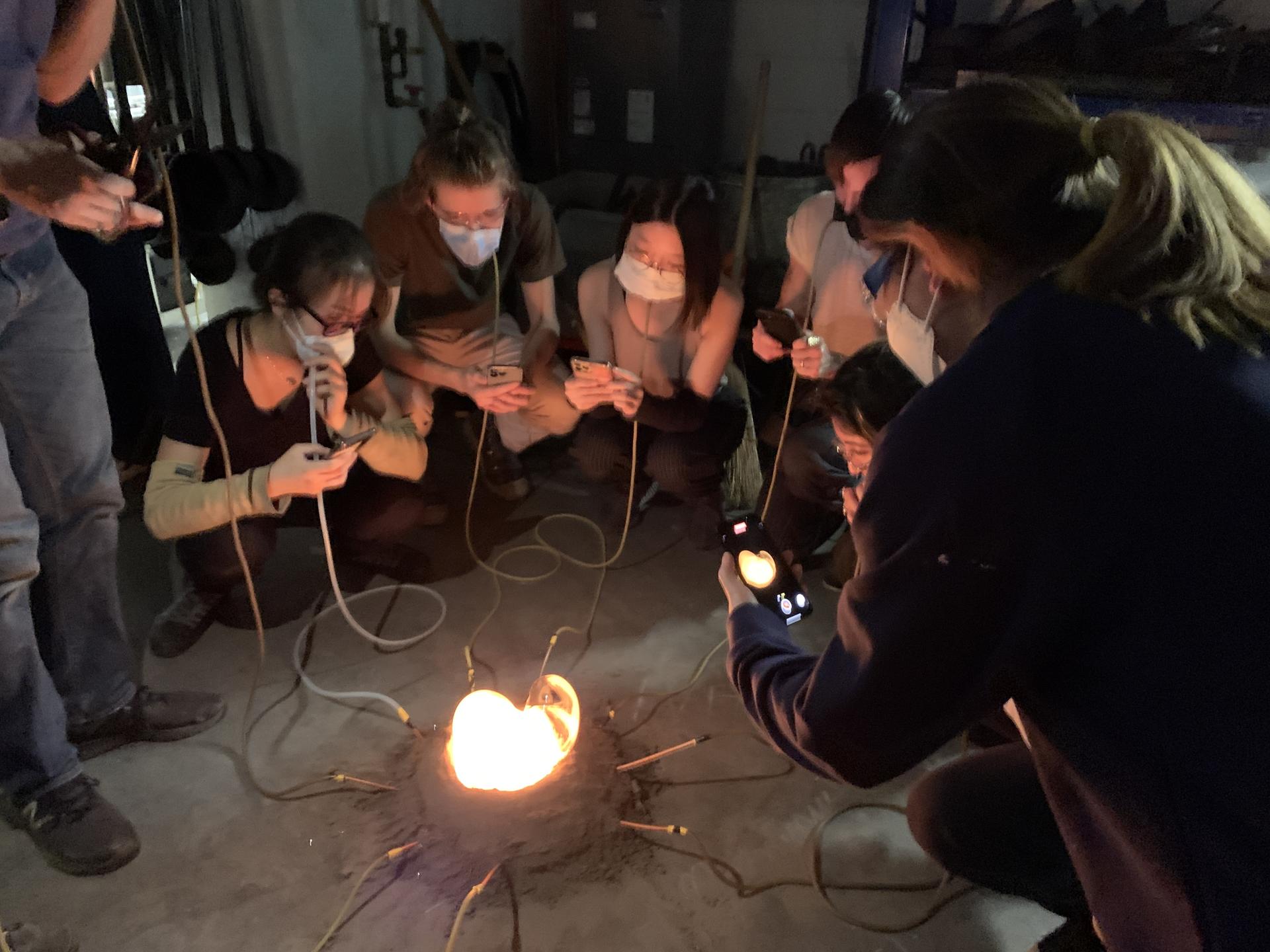 A group of students surround a glowing lump of glass in a pile of dirt on the floor. Each has a tube in their mouth that connects to the pile of dirt on the floor below the glass.