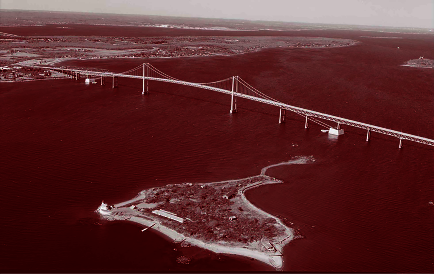 red-tinted aerial view of the Pell Bridge, a suspension bridge spanning a river with an island 