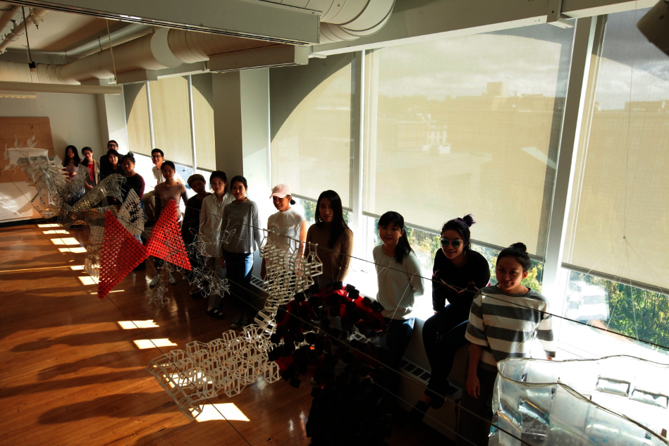 A row of students sit with 3D work in front of windows