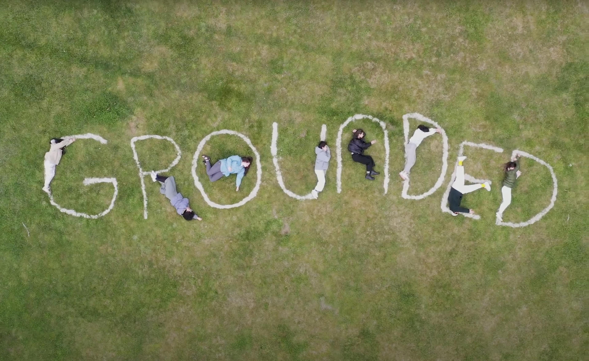 Aerial photo of a field of grass with the word "grounded" spelled out. Human figures lay on the ground, interacting with the letters.