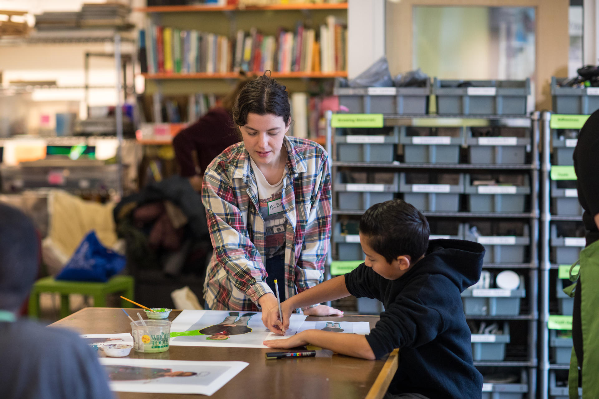 In a classroom setting, a young white woman assists a Latinx child holding a pencil as he draws on a photograph.