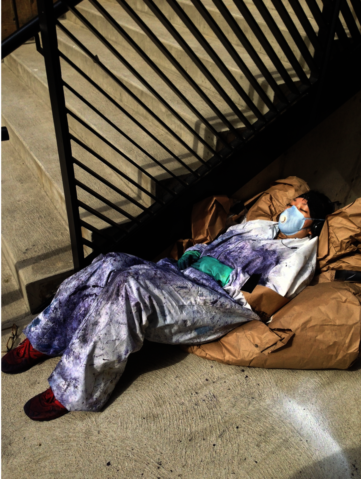 A tired student reclines on brown bags by stairs with a metal railing. They are wearing headphones, rubber gloves, a dye-splattered lab coat, and a respirator mask.