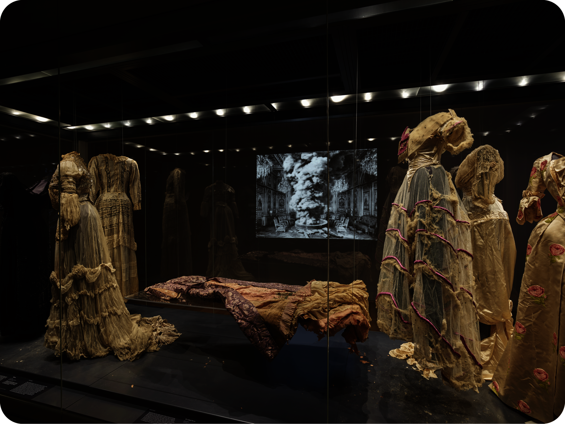 View of several ornate cream-colored dresses on mannequins encased in glass. A table in the center holds another dress. In the background is a projection of a grand ornate space. 